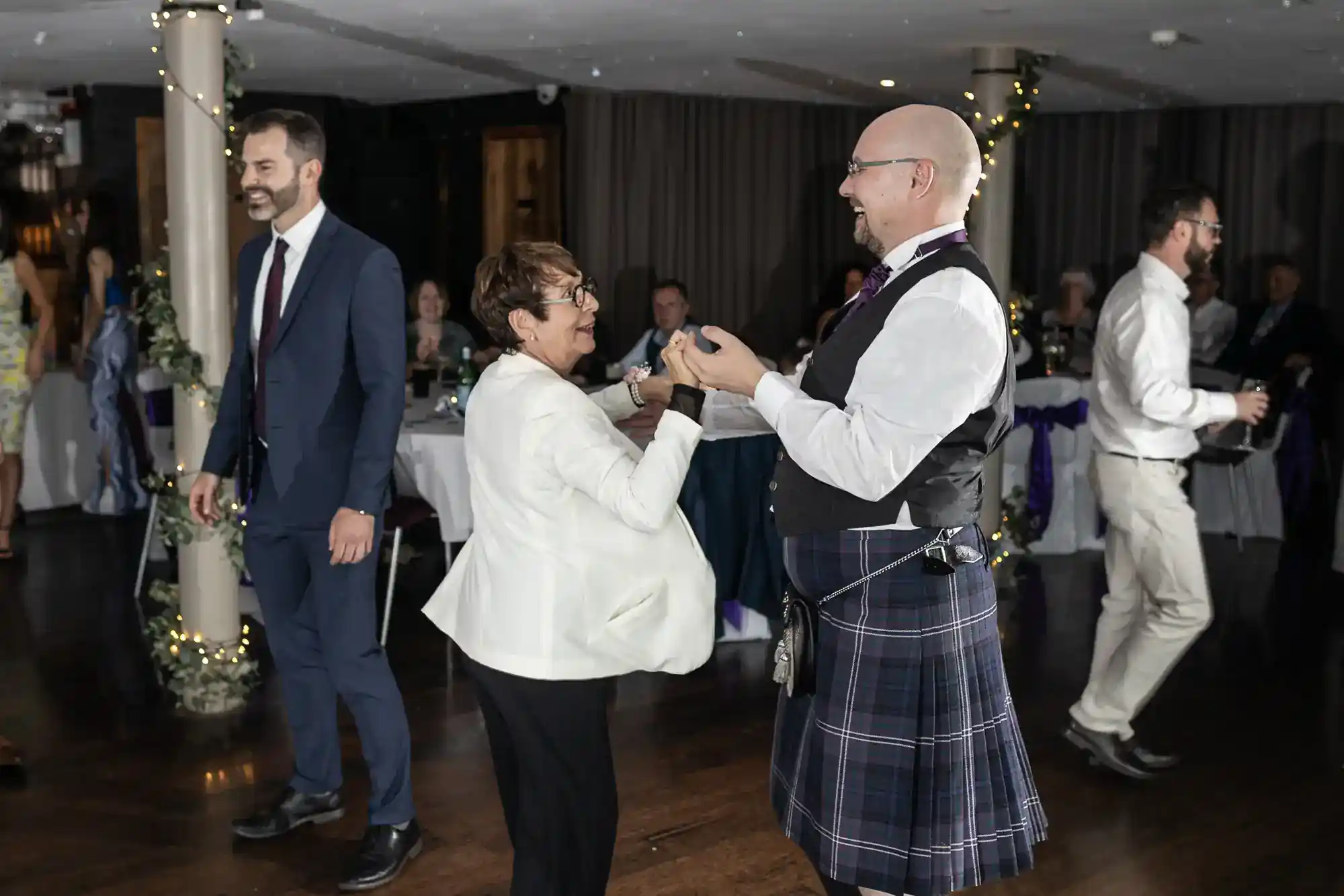 An older woman and a man in a kilt, waistcoat, and glasses dance together at a wedding reception. Other guests stand and mingle in the decorated room, with tables in the background and fairy lights wrapped around a pillar.