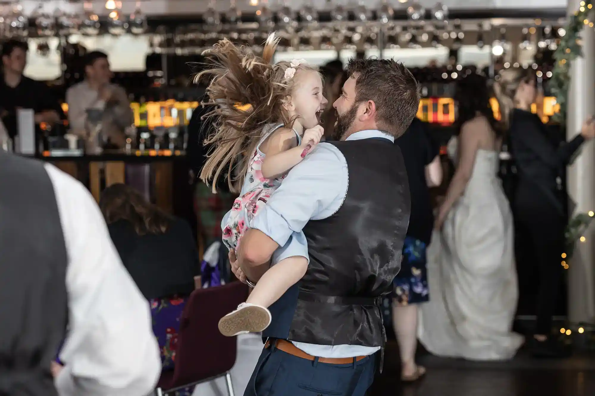 A man in a waistcoat spins a laughing young girl in a floral dress at a lively indoor event; other guests, including a woman in a white dress, socialise and dance in the blurred background under fairy lights.