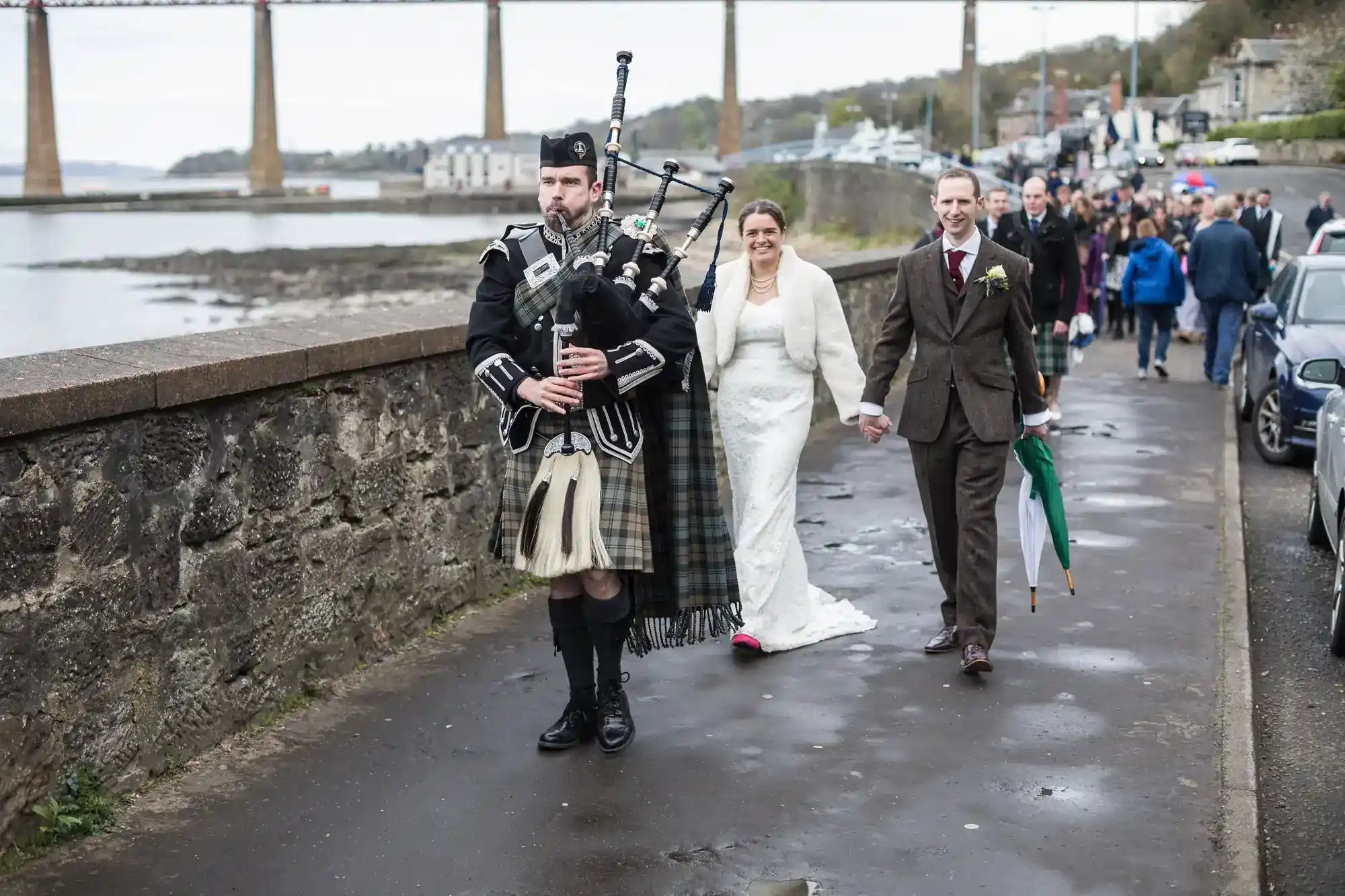A bagpiper in traditional Scottish attire leads a bride in a white dress and a groom in a suit, holding hands and smiling, along a riverside path. Wedding guests walk behind them. A large bridge and water are visible in the background.