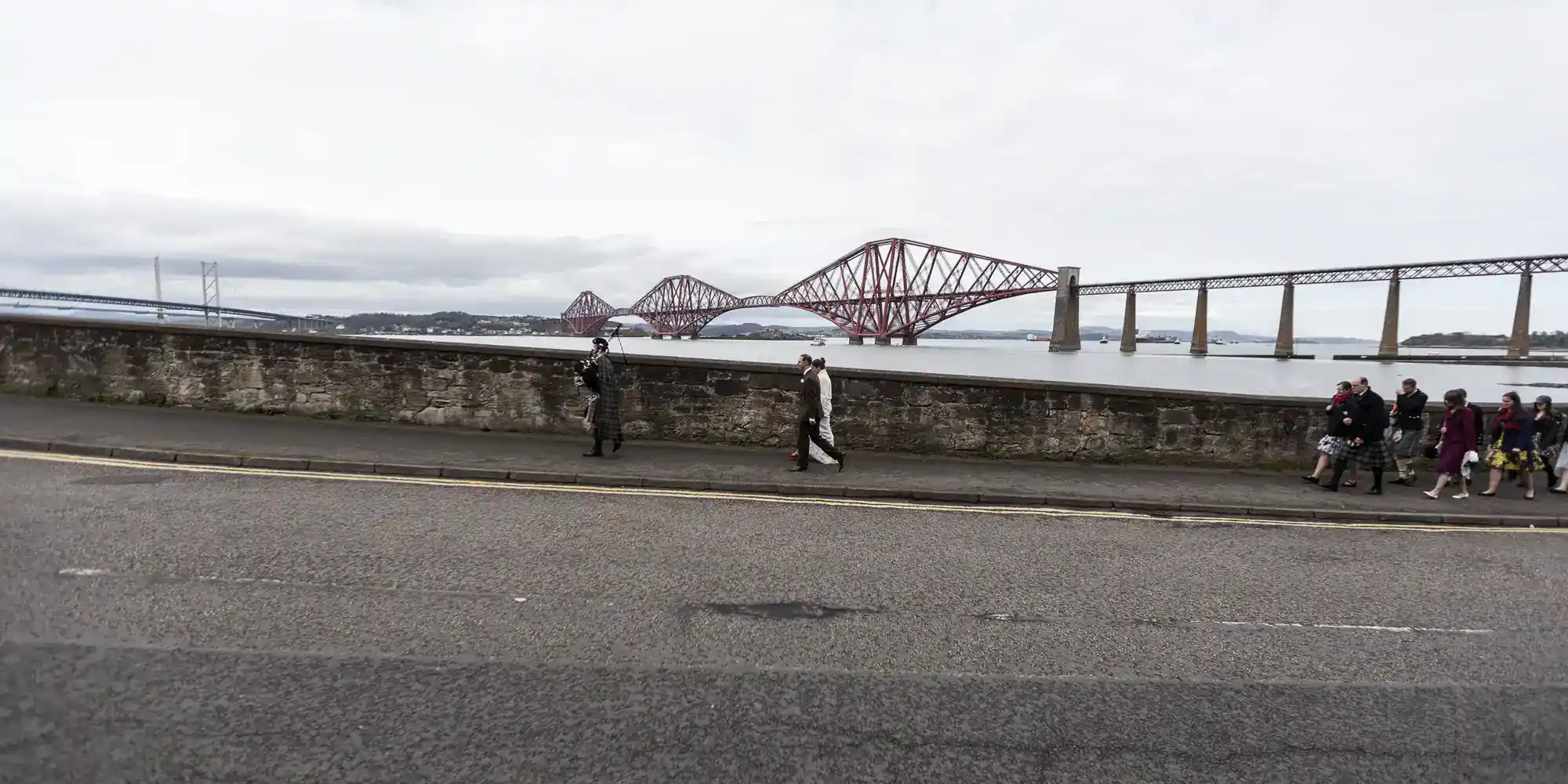A group of people, some in fancy dress, walk along a pavement by a stone wall. Behind them is water and a large red cantilever railway bridge. The sky is overcast and road markings are visible in the foreground.