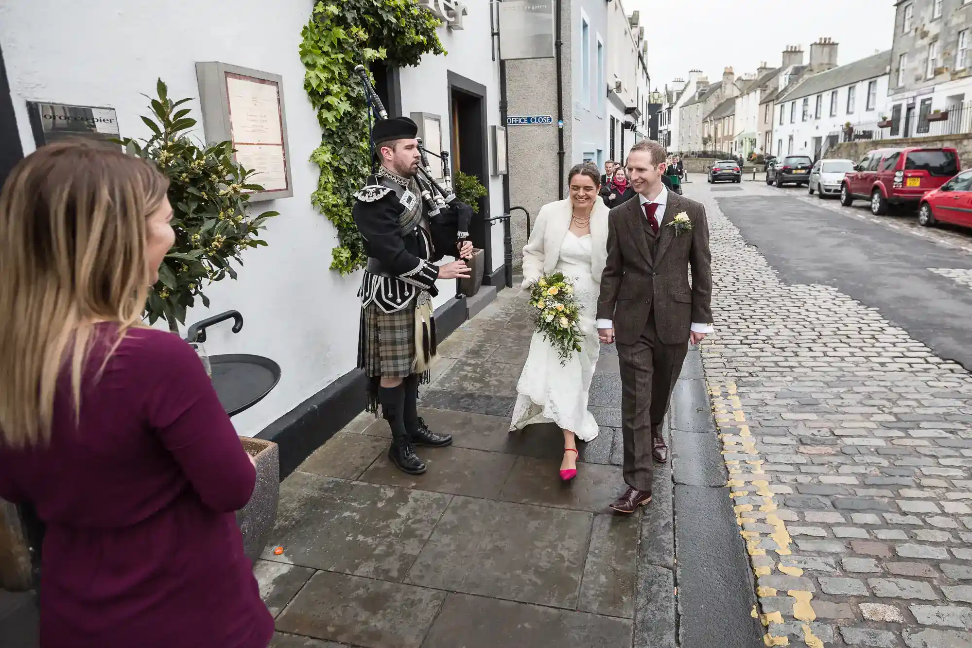 A bride and groom walk hand-in-hand down a cobbled street, smiling. A bagpiper in traditional Scottish attire plays nearby whilst a woman in a burgundy dress watches. The scene is festive, set outside on a cloudy day in a quaint town.