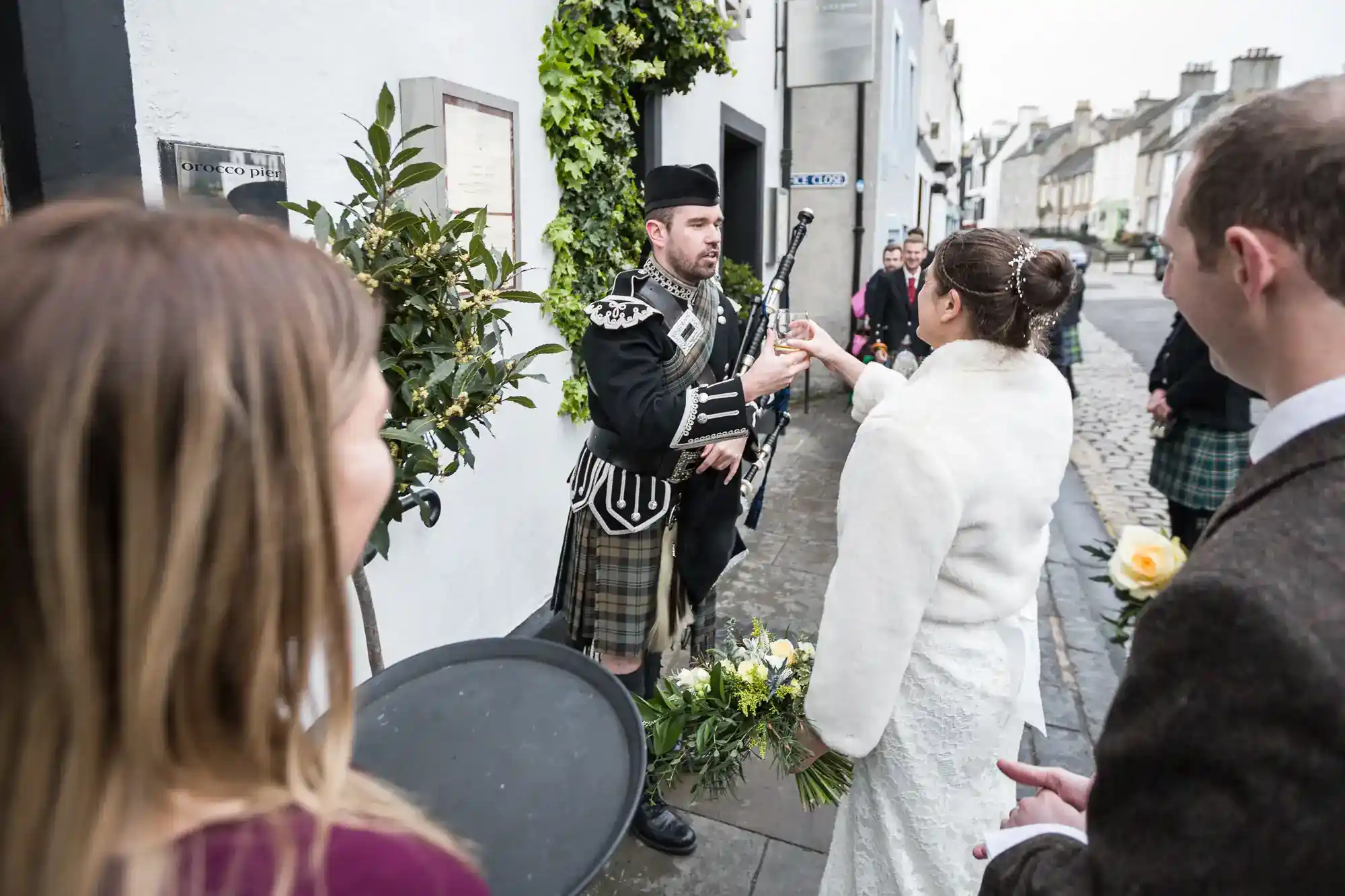 A bride in a white coat hands a glass to a man in traditional Scottish attire playing bagpipes on a stone street. Other people, some holding flowers, stand nearby, and the scene is festive with greenery and historic buildings in the background.
