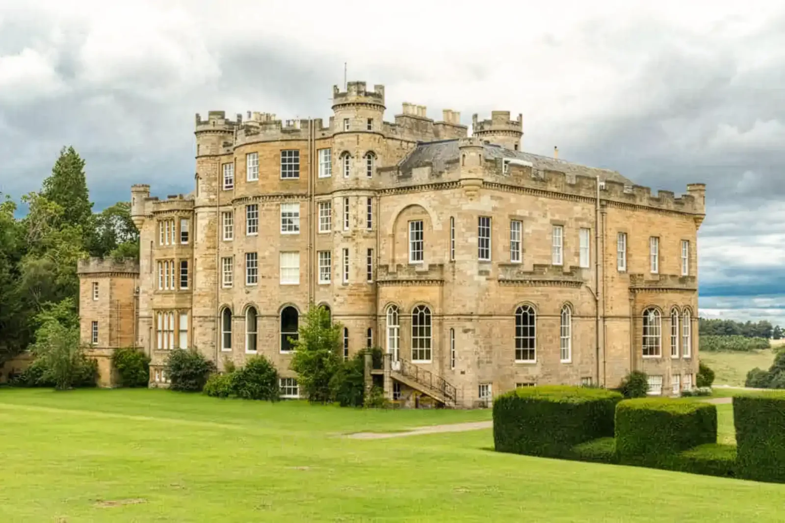 A large, historic stone Oxenfoord Castle with multiple arched windows, corner turrets, and battlements stands on a manicured green lawn, bordered by trimmed hedges and trees, beneath a cloudy sky.
