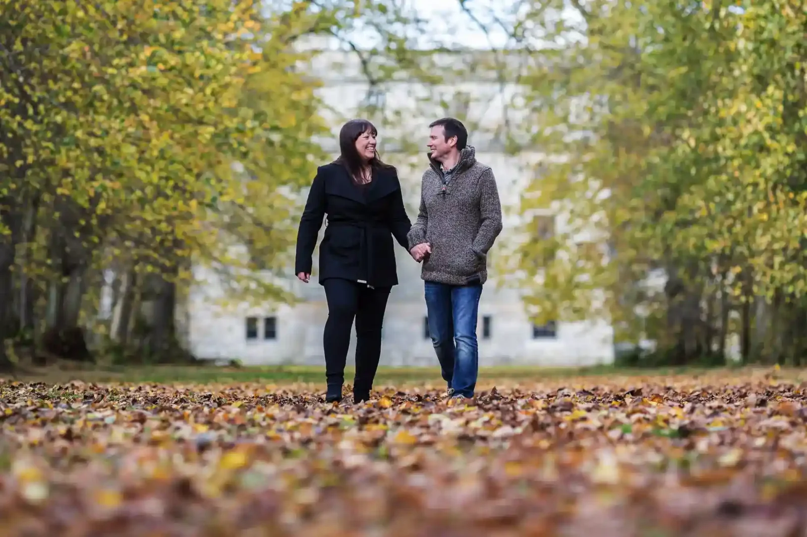A couple walks hand in hand through Penicuik Estate covered in autumn leaves. They smile at each other, wearing jackets and jeans. Trees with yellow and green foliage surround them. A blurred large building is visible in the background, blending with the scene.