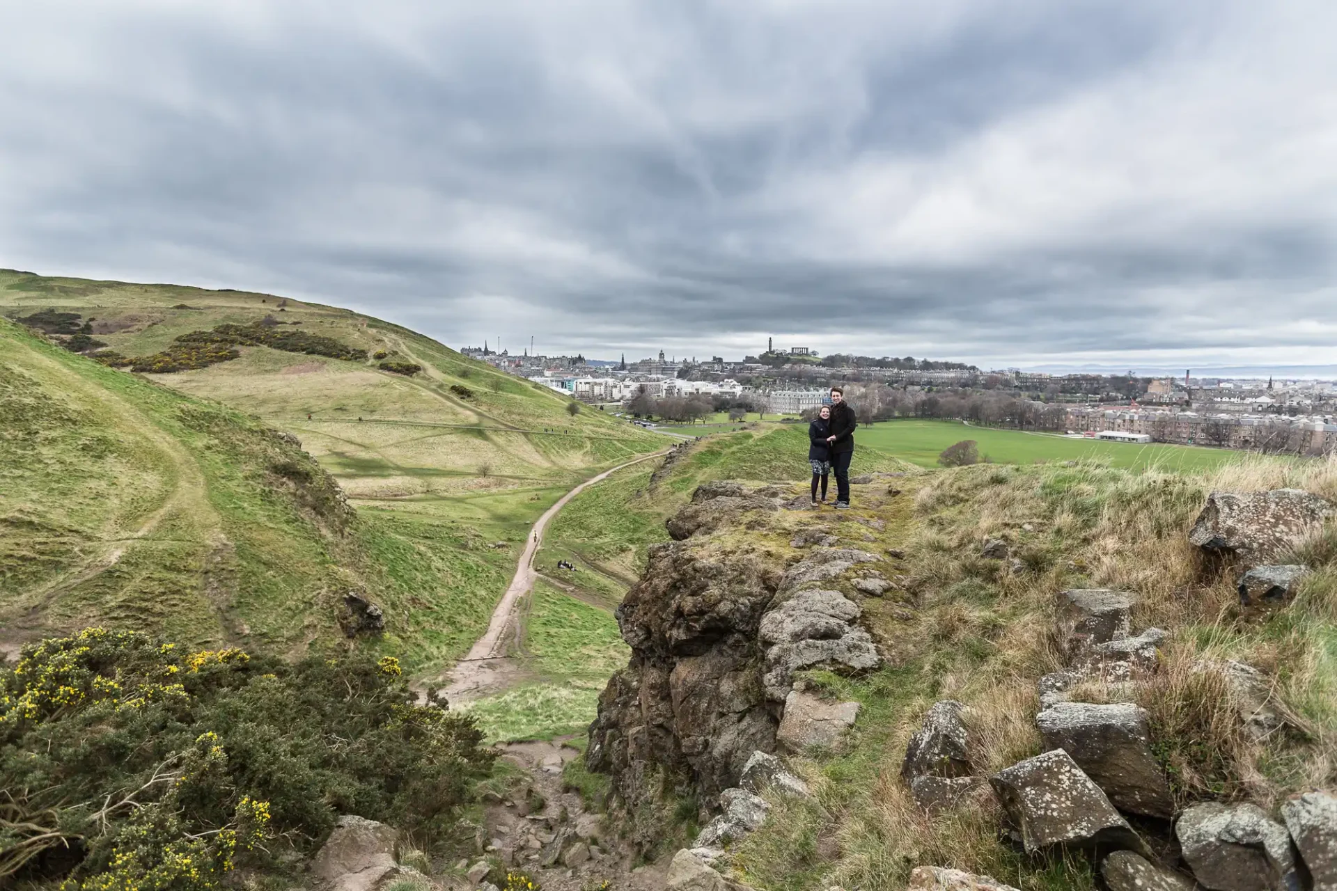 Engagement photos session at Arthur's Seat - Jon and Mandy: Two people stand on a rocky outcrop overlooking a vast grassy landscape with a distant city skyline under a cloudy sky.
