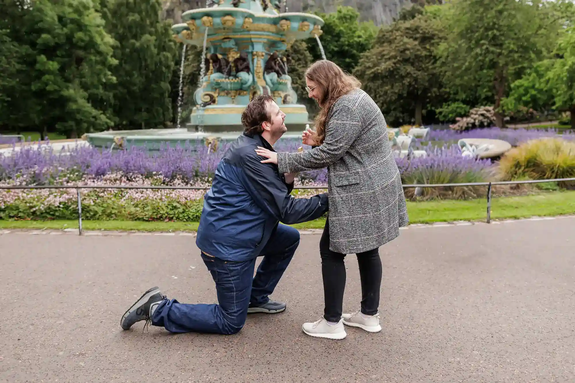 A man kneels and proposes to a surprised woman in front of a decorative fountain in a park, Ross Fountain in Princes Street Gardens in Edinburgh, surrounded by blooming purple flowers and lush green trees. Both are dressed in casual, warm clothing and appear happy and emotional, the moment captured by proposal photographer in Edinburgh Jonathon Fowler Photography.