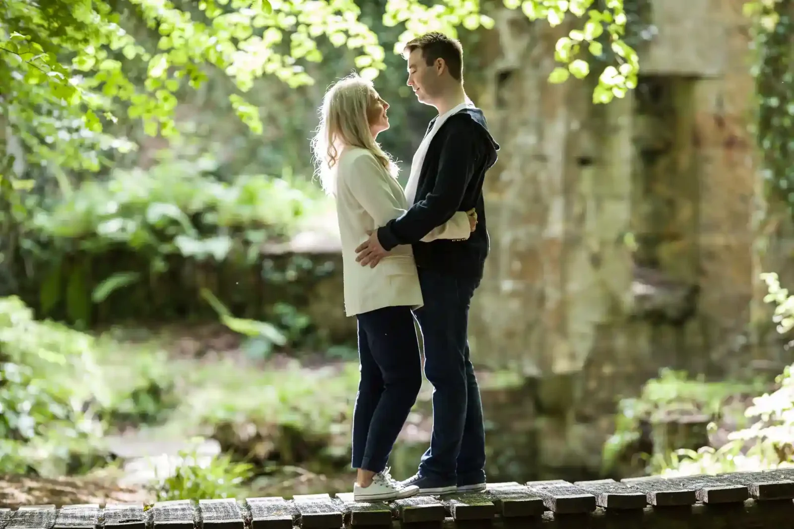 A smiling couple stands face to face on a wooden bridge in a sunlit, green Roslin Glen. The woman wears a white top and jeans; the man wears a dark jacket and jeans. They hold each other, surrounded by trees and foliage, with soft natural light overhead.