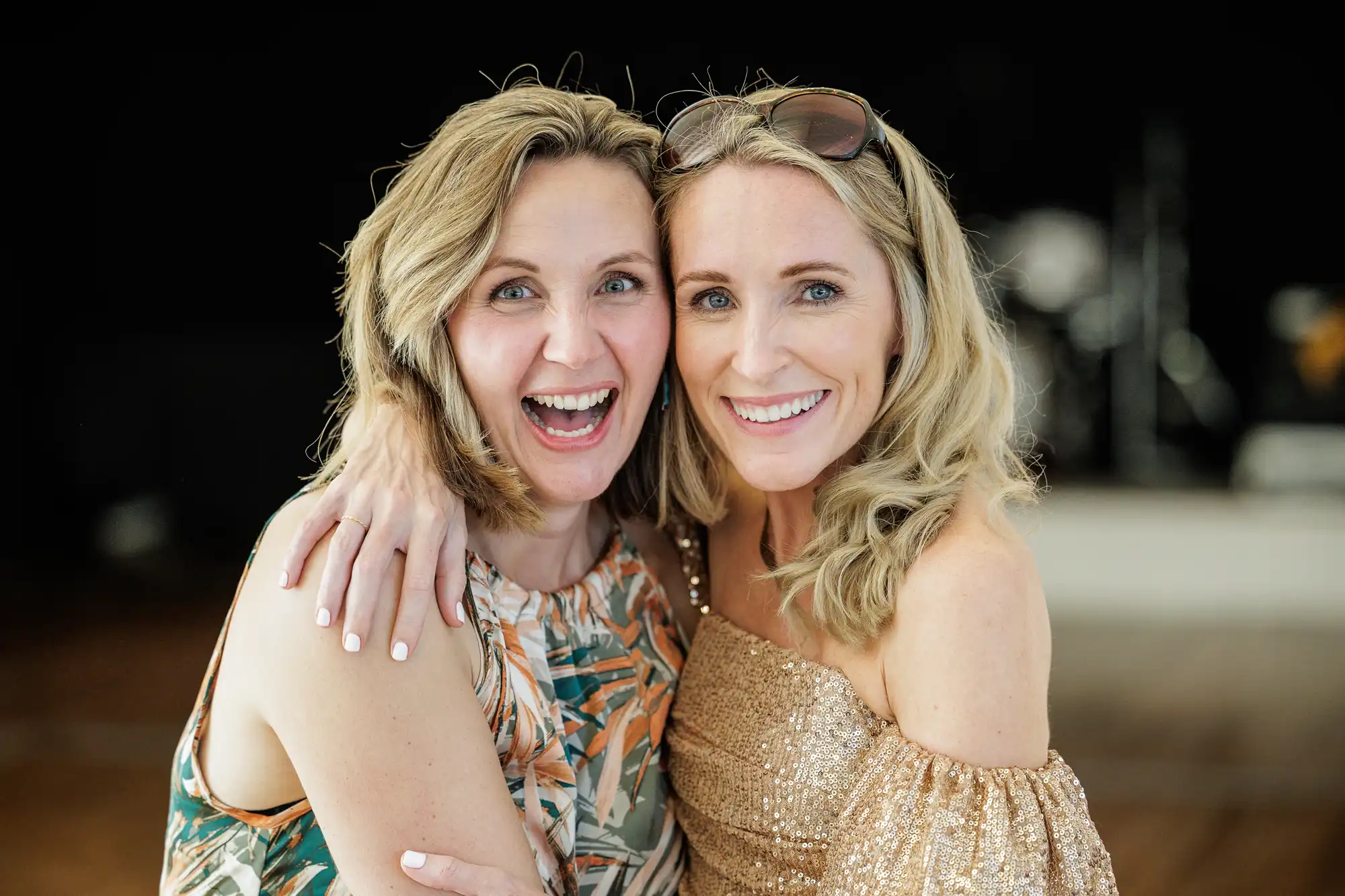 Runningburn wedding photographer image of two smiling women with blonde hair pose closely together, one wearing a patterned sleeveless dress and the other in a sparkly off-the-shoulder top with sunglasses on her head. They stand indoors against a blurred dark background.