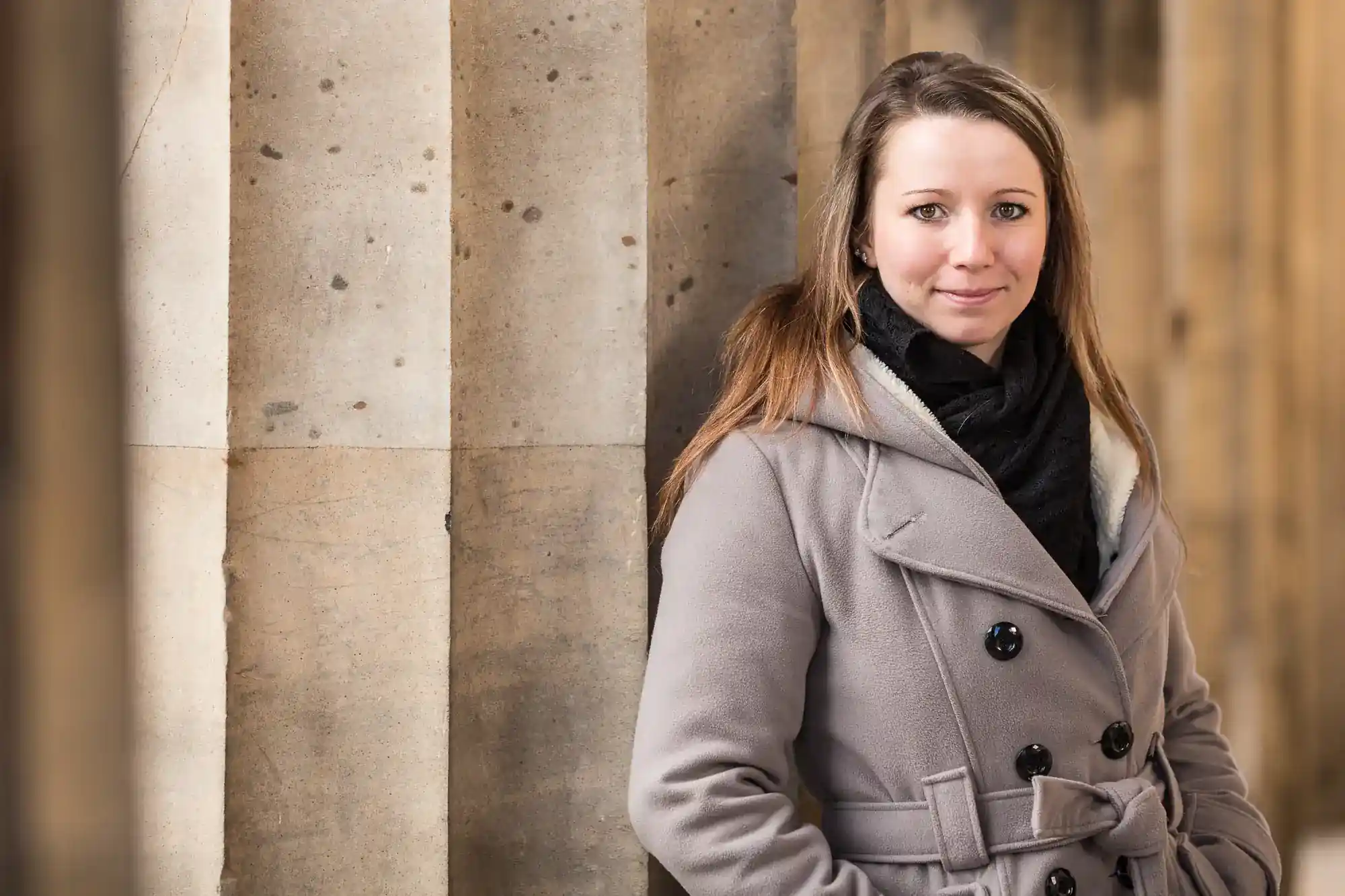 A woman with long light brown hair, wearing a grey coat and black scarf, stands beside large stone columns. She is slightly smiling and looking at the camera. The background is softly blurred, highlighting the beige columns and her relaxed posture.