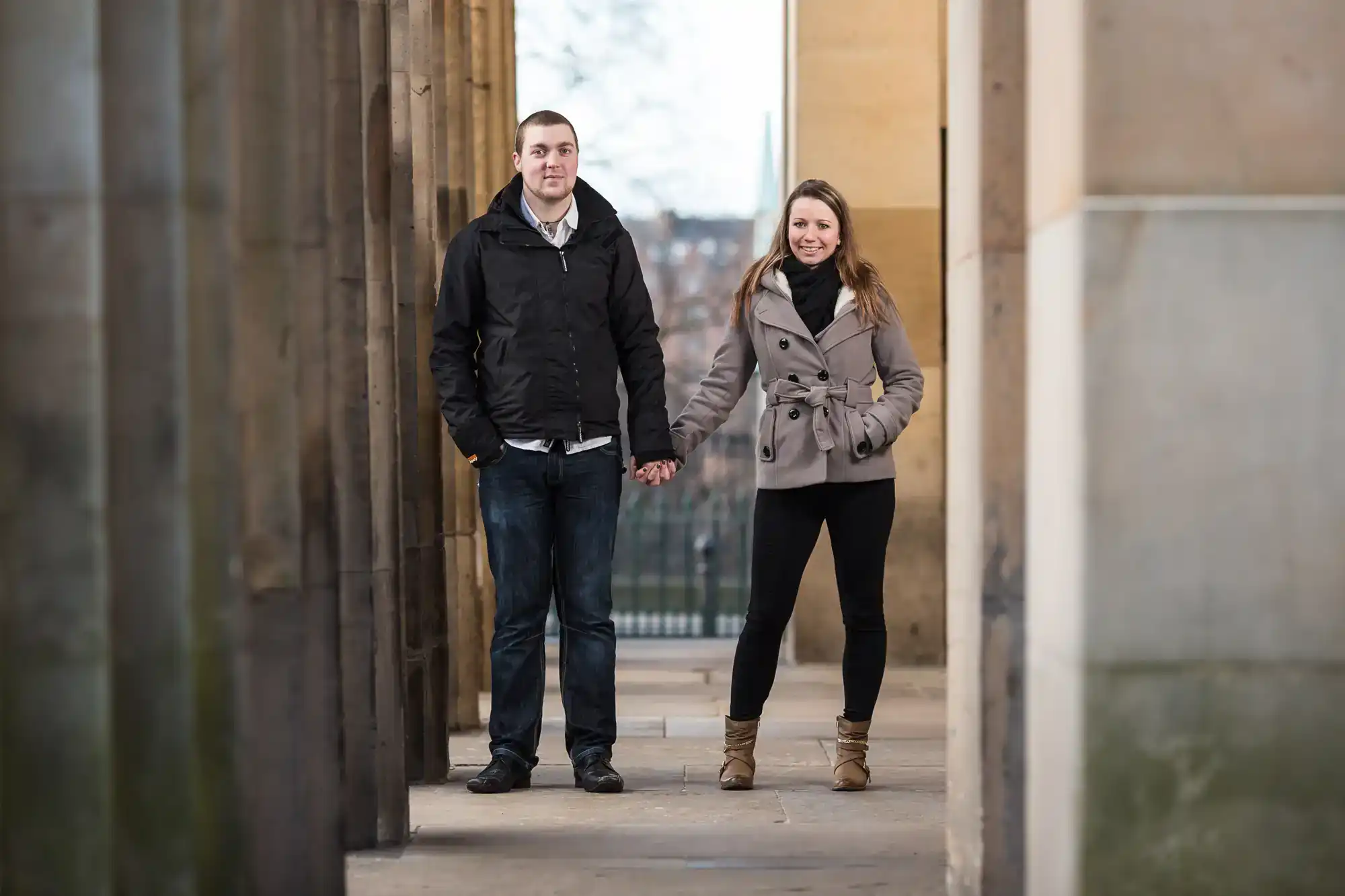 A young man and woman stand holding hands between tall stone columns. Both wear jackets, jeans, and boots. They face the camera and smile slightly. The background shows an outdoor urban setting with blurred trees and buildings visible.