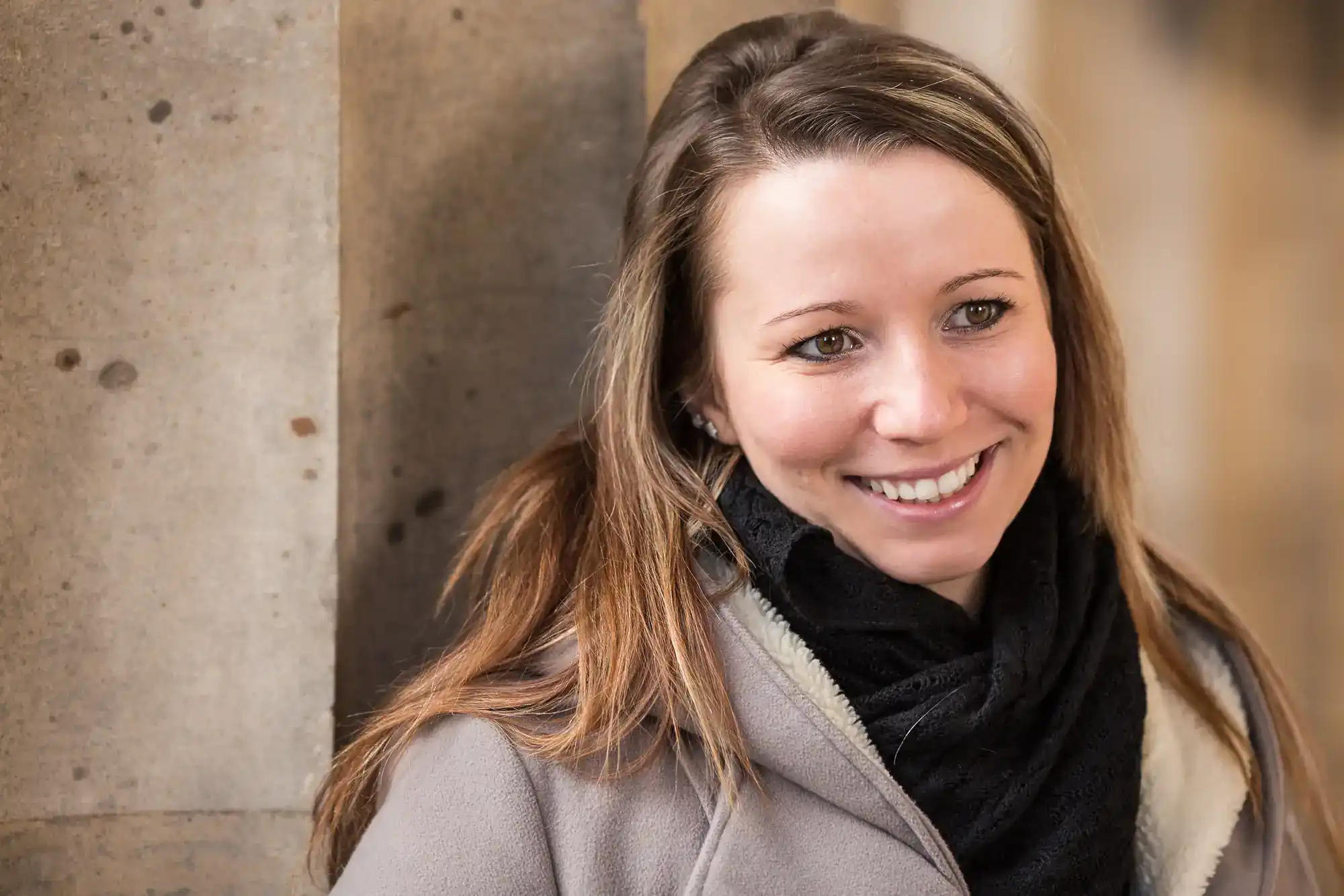 A woman with straight, light brown hair and a black scarf smiles whilst leaning against a stone wall. She wears a light grey coat and looks slightly to the right. The background is softly blurred, emphasising her face and friendly expression.