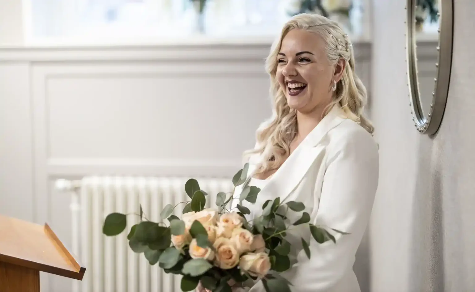 A smiling woman with long, wavy blonde hair wearing a white outfit holds a bouquet of pale peach roses and green leaves. She stands indoors near a wooden lectern, in front of a radiator and a wall mirror.