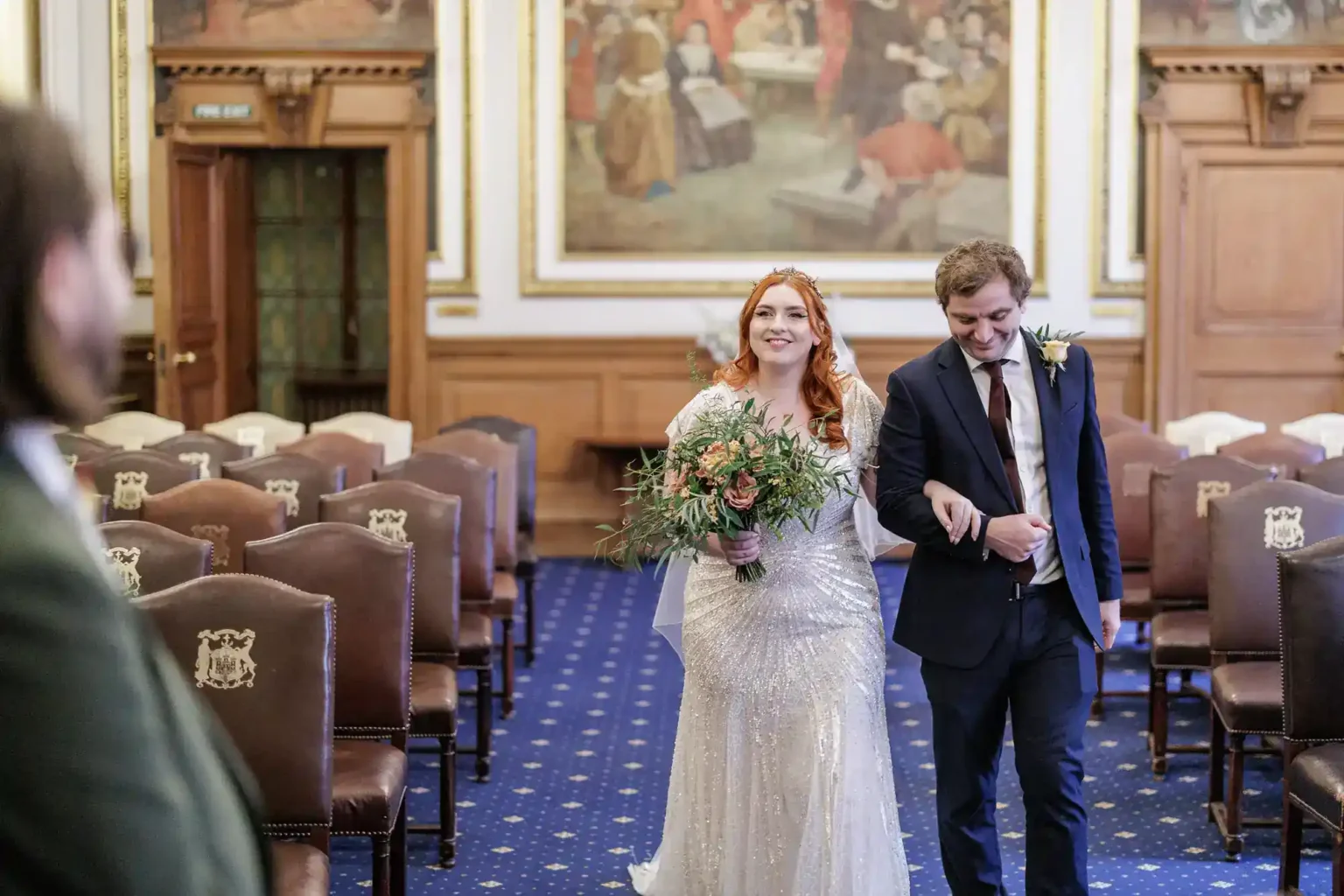 A bride in a sparkly white dress holding a bouquet and a smiling groom walk arm-in-arm down the aisle of an ornate, empty hall, with brown chairs, blue carpet, and large paintings on the wall. Another person stands blurred in the foreground, watching them.