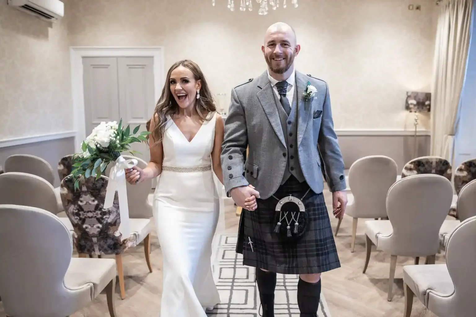 A smiling bride in a white dress holds a bouquet and the hand of a groom in a grey jacket, kilt, and sporran. They walk down an aisle in a light, elegant room with rows of beige chairs, celebrating their wedding.