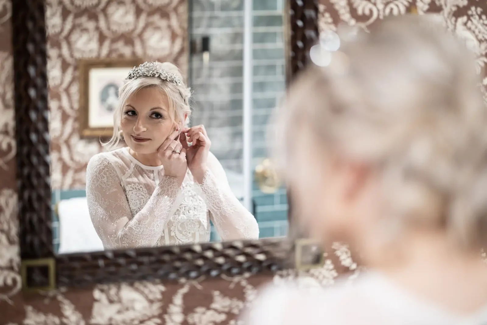 A bride with blonde hair and a jewelled tiara puts on earrings while looking in a mirror. She wears a white lace wedding dress. The ornate mirror reflects her focused expression. The background features patterned wallpaper and a framed photo.