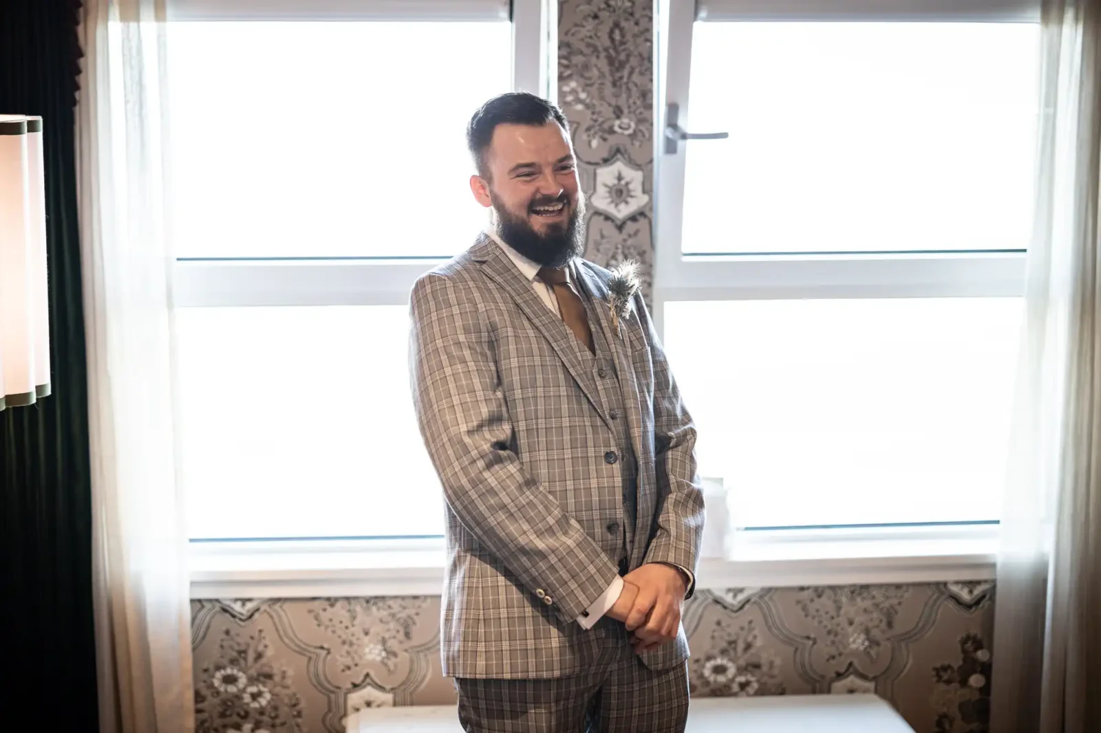 A bearded man in a grey checked suit smiles warmly whilst standing indoors in front of two bright windows with white curtains. Ornate patterned wallpaper and soft, diffused light fill the room, creating a cosy and elegant atmosphere.