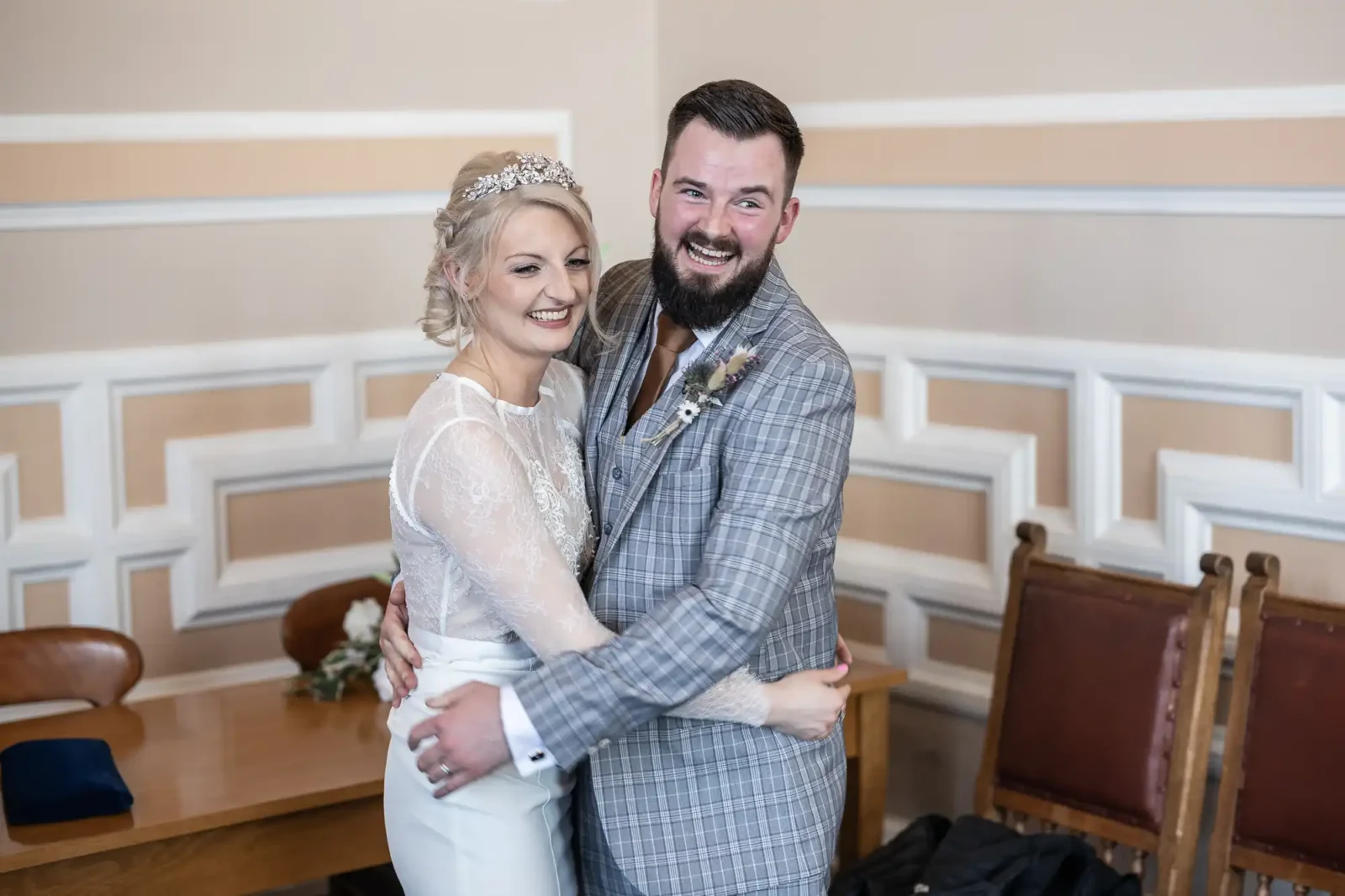 A bride in a white lace dress and tiara embraces a groom in a grey checked suit with a buttonhole. Both are smiling joyfully. They stand in a room with cream walls, wooden chairs, and a table in the background.