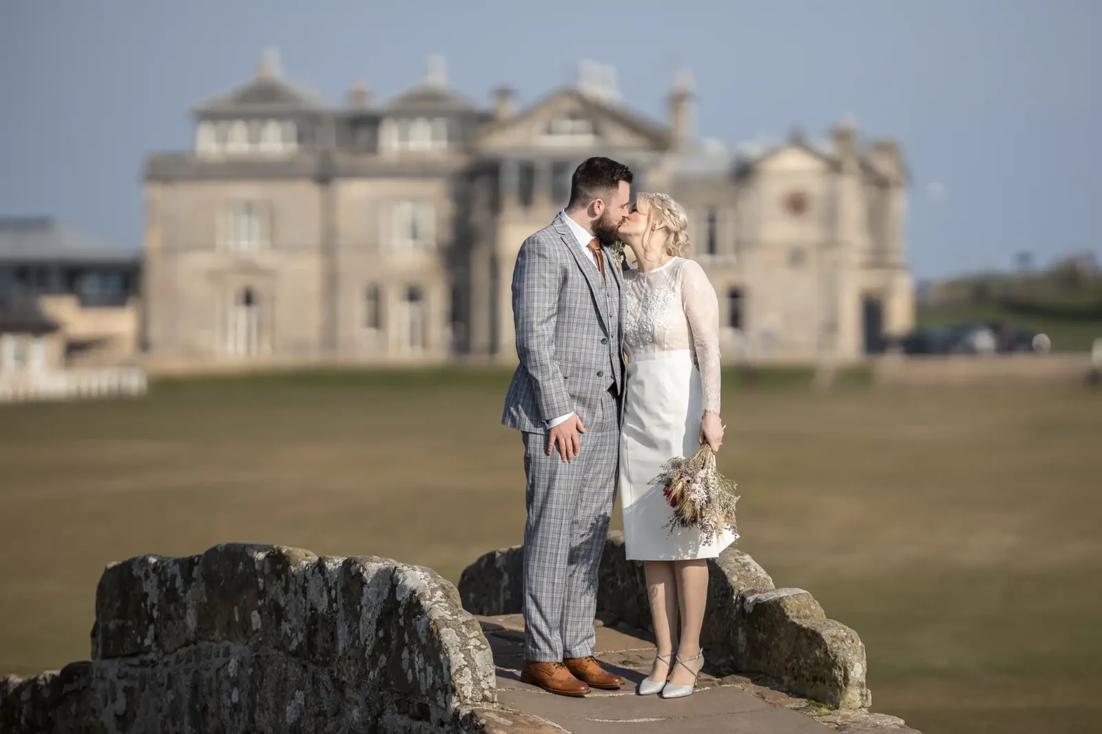 A bride and groom kiss on a stone bridge. The groom wears a grey checked suit, whilst the bride wears a white knee-length dress and holds a bouquet. A large historic building with many windows is blurred in the background under a clear sky.