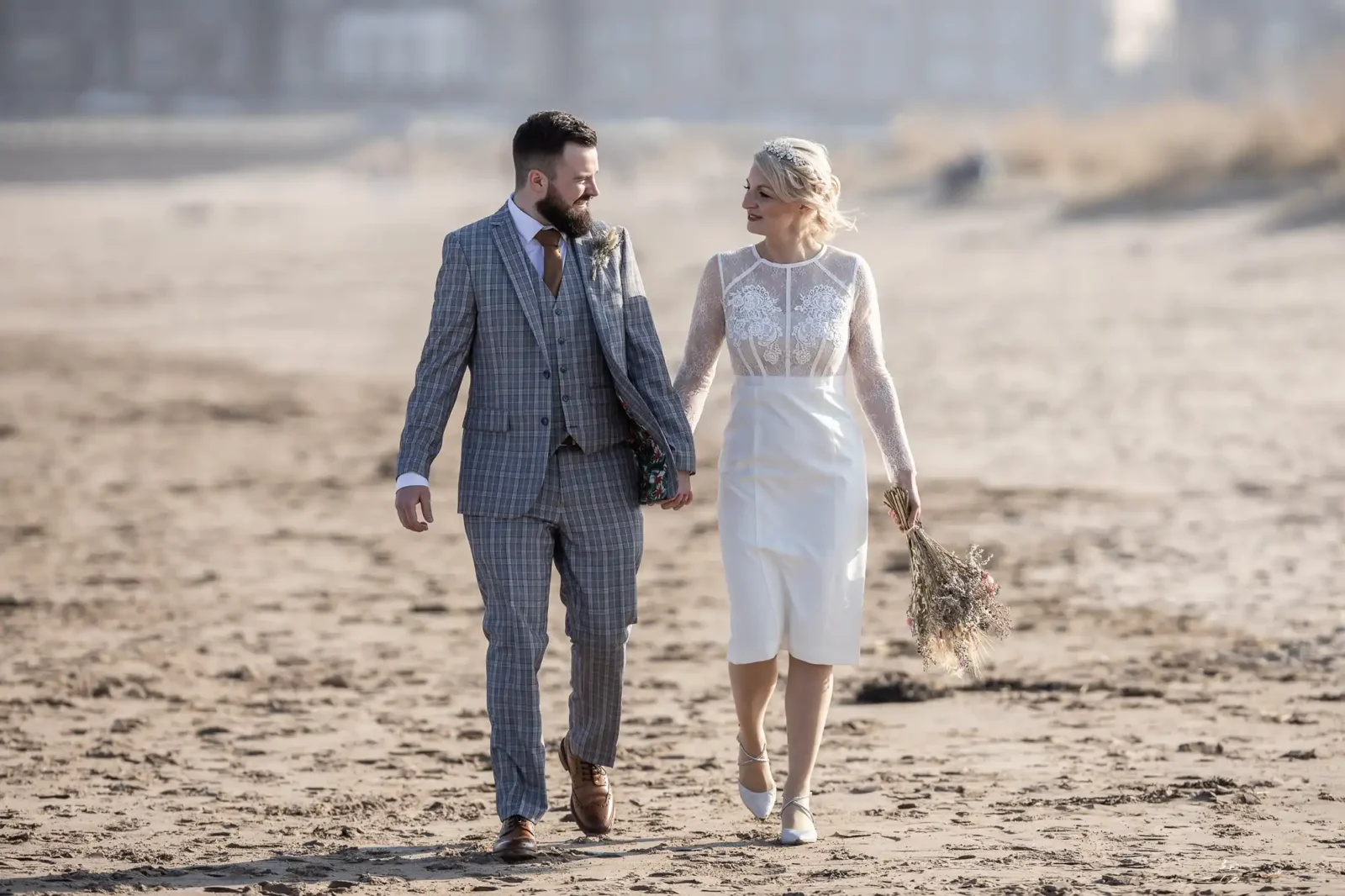 A couple in wedding attire walk barefoot on a sandy beach. The groom wears a grey checked suit and brown shoes, while the bride wears a white lace dress and carries a bouquet of dried flowers. They are holding hands, looking at each other, with soft sunlight and blurred background.