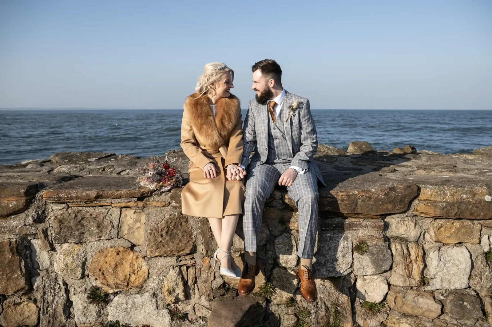 A couple dressed in formal attire sit on a stone wall by the sea, holding hands and smiling at each other. The woman wears a tan coat with a fur collar and silver heels; the man wears a grey checked suit with a brown tie. A bouquet rests beside them.