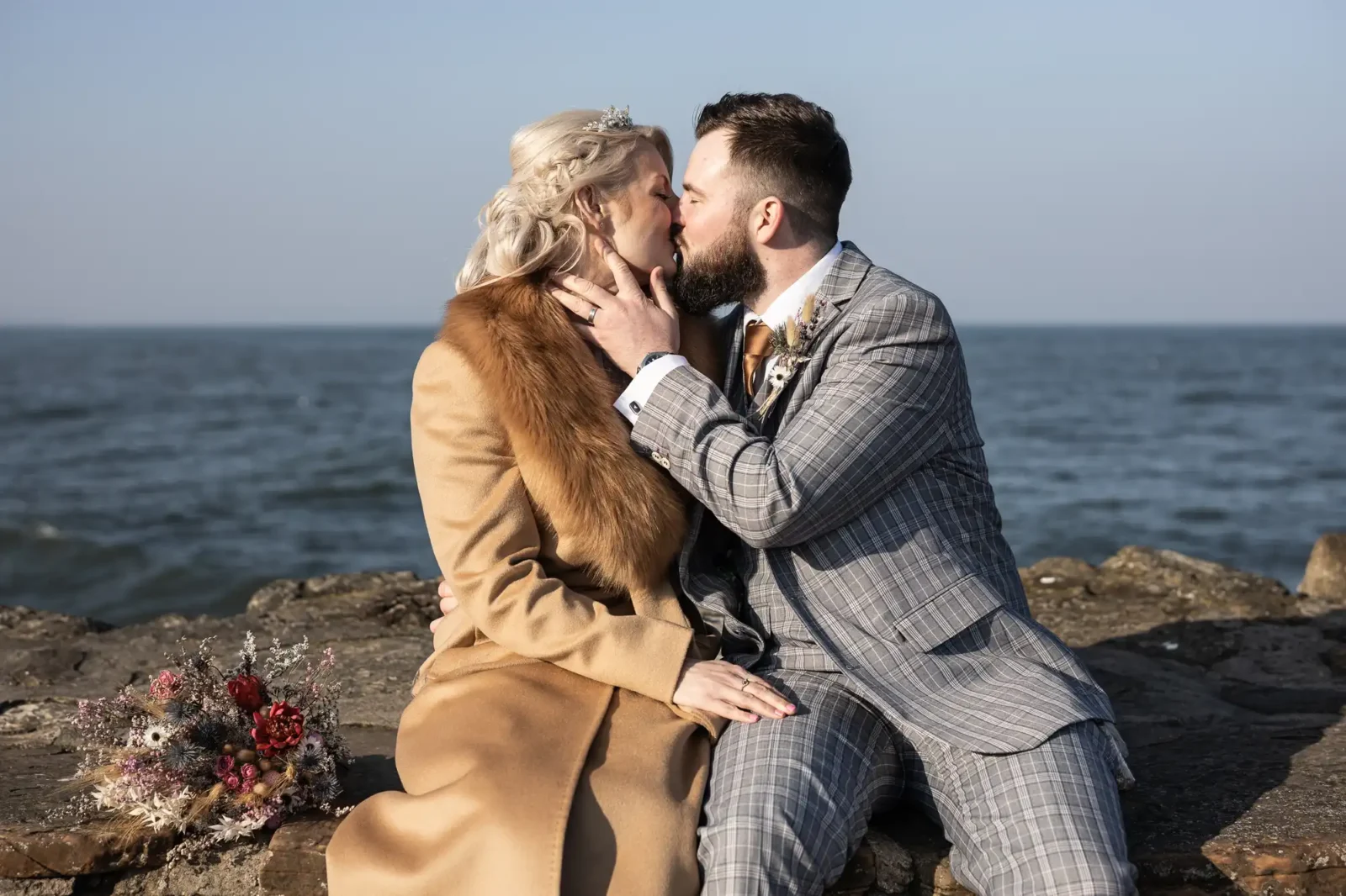A couple in formal attire sits on a stone wall by the sea, kissing. The woman wears a tan coat with a fur collar; the man wears a grey checked suit. A bouquet of flowers rests beside them. The sky is clear and blue.