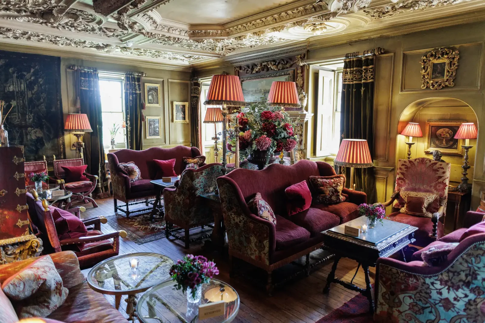 Ornate vintage living room with carved ceiling, tall windows, antique furniture, red velvet sofas, floral armchairs, patterned rugs, lamps with red shades, bouquets of flowers on tables, and classic artwork on the walls. Warm, cosy and richly decorated.