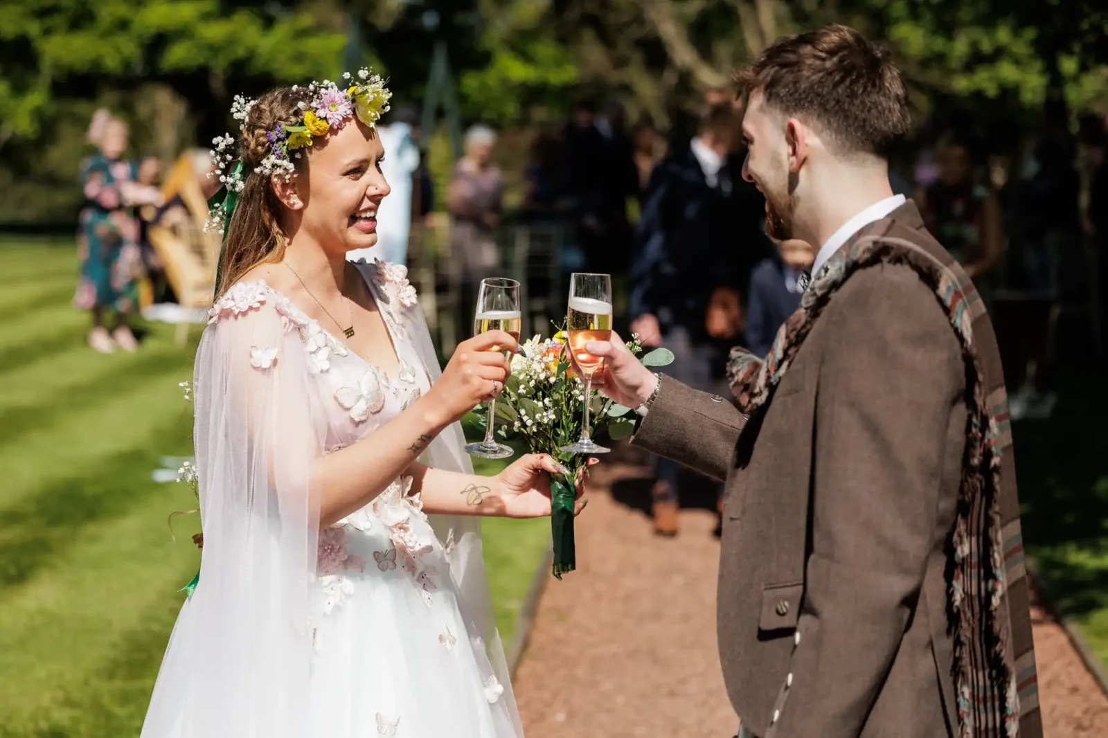 A bride in a white floral dress and flower crown smiles as she toasts with a groom in a brown suit. They hold champagne glasses and a bouquet, standing outdoors on a sunny day with green grass and guests in the background.