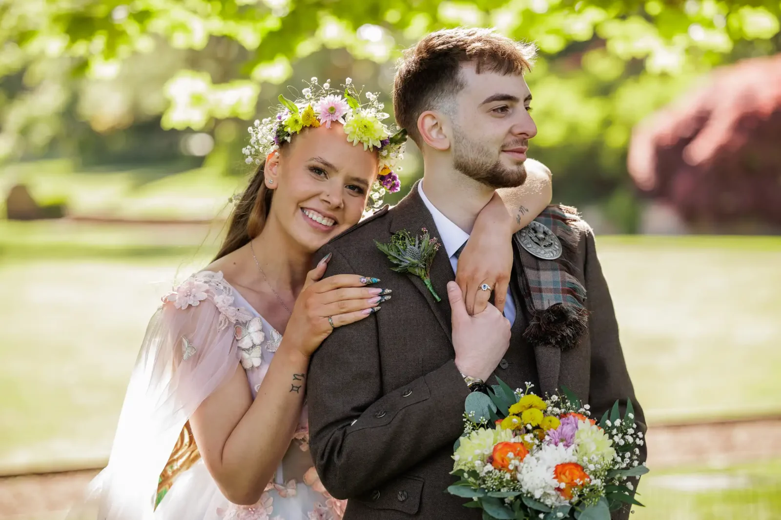 A summer wedding at Prestonfield House showing a smiling bride with a floral crown embraces the groom from behind, resting her head on his shoulder. The groom, in a brown suit with a checked sash, holds a colourful bouquet. They stand outdoors in sunlight, with greenery blurred in the background.