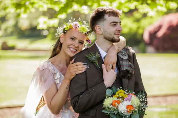 A summer wedding at Prestonfield House showing a smiling bride with a floral crown embraces the groom from behind, resting her head on his shoulder. The groom, in a brown suit with a checked sash, holds a colourful bouquet. They stand outdoors in sunlight, with greenery blurred in the background.