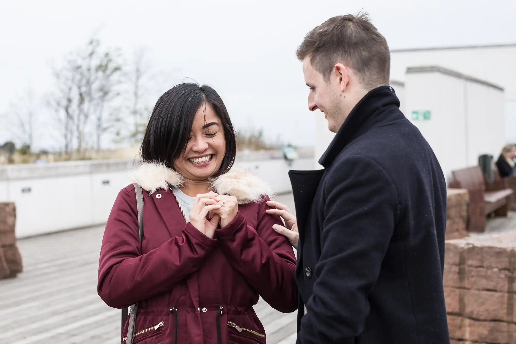 A woman and a man holding hands and smiling at each other outdoors, both wearing coats.