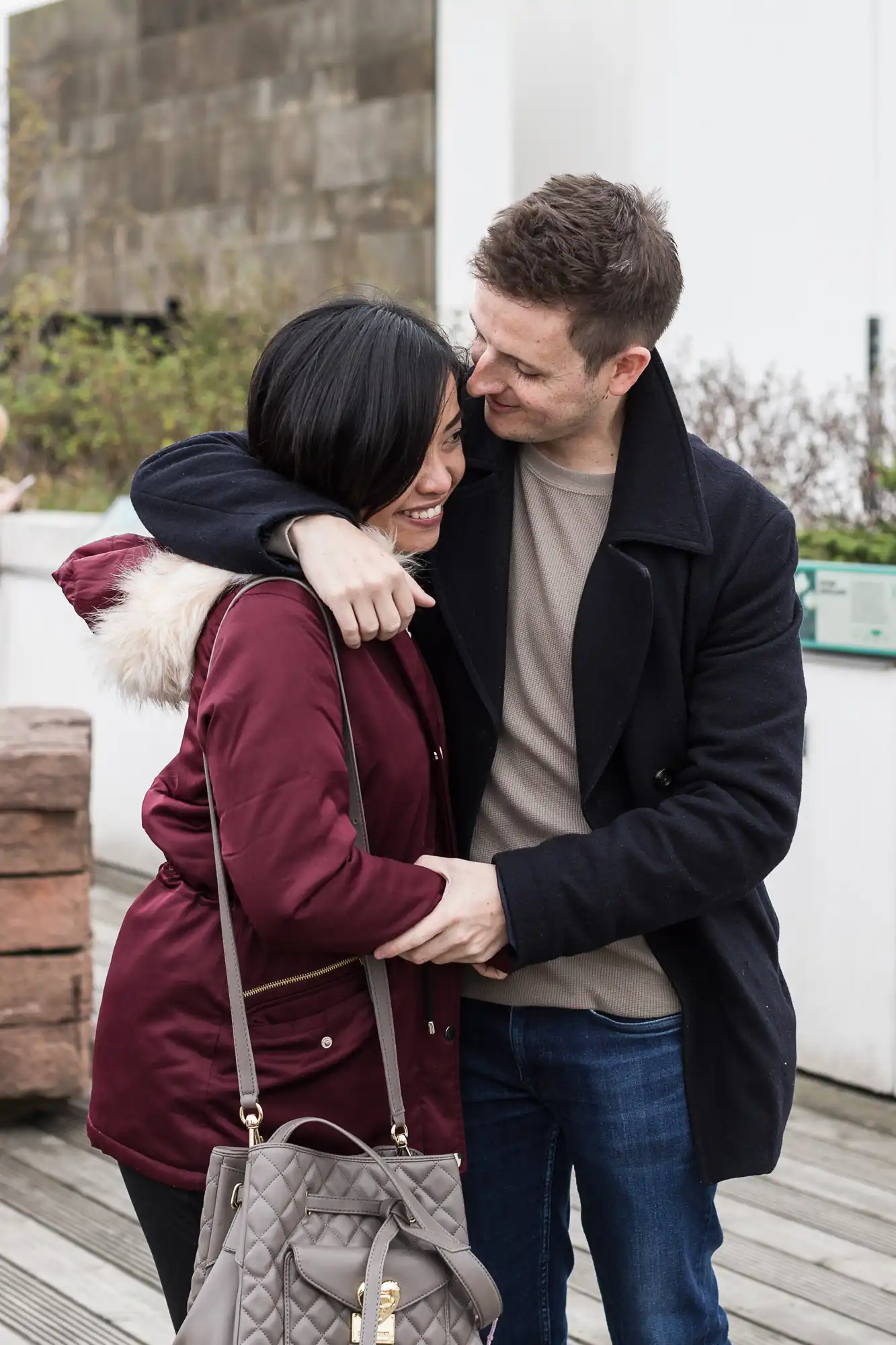 A couple embracing and smiling on a wooden deck, man in a coat and woman in a red jacket, both looking happy.