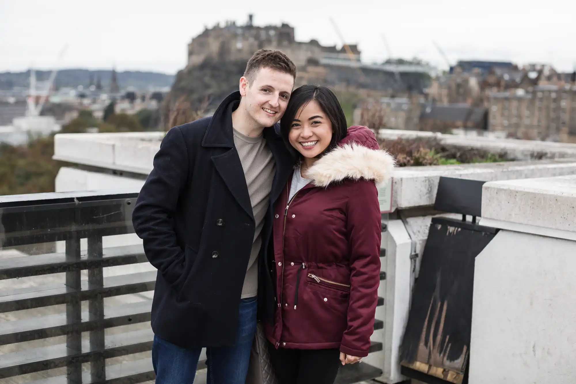 A couple embracing and smiling at the camera with a historical castle in the background on a cloudy day.
