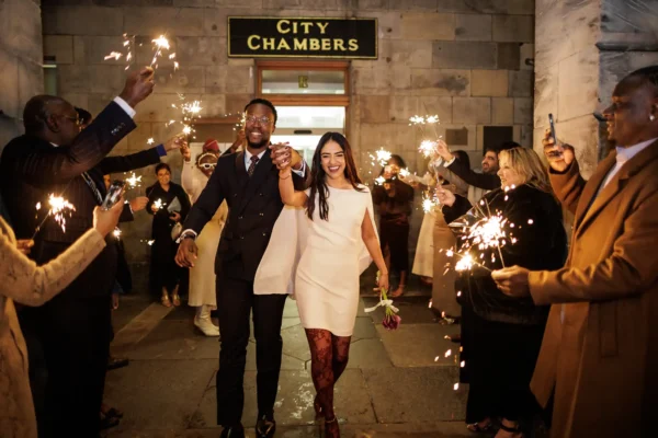 The Alexander Suite winter wedding photo of a joyful couple walk hand-in-hand outside City Chambers, smiling as they are surrounded by well-dressed guests holding sparklers. The couple are dressed formally; the woman holds a bouquet and wears a white dress, whilst the man wears a dark suit.