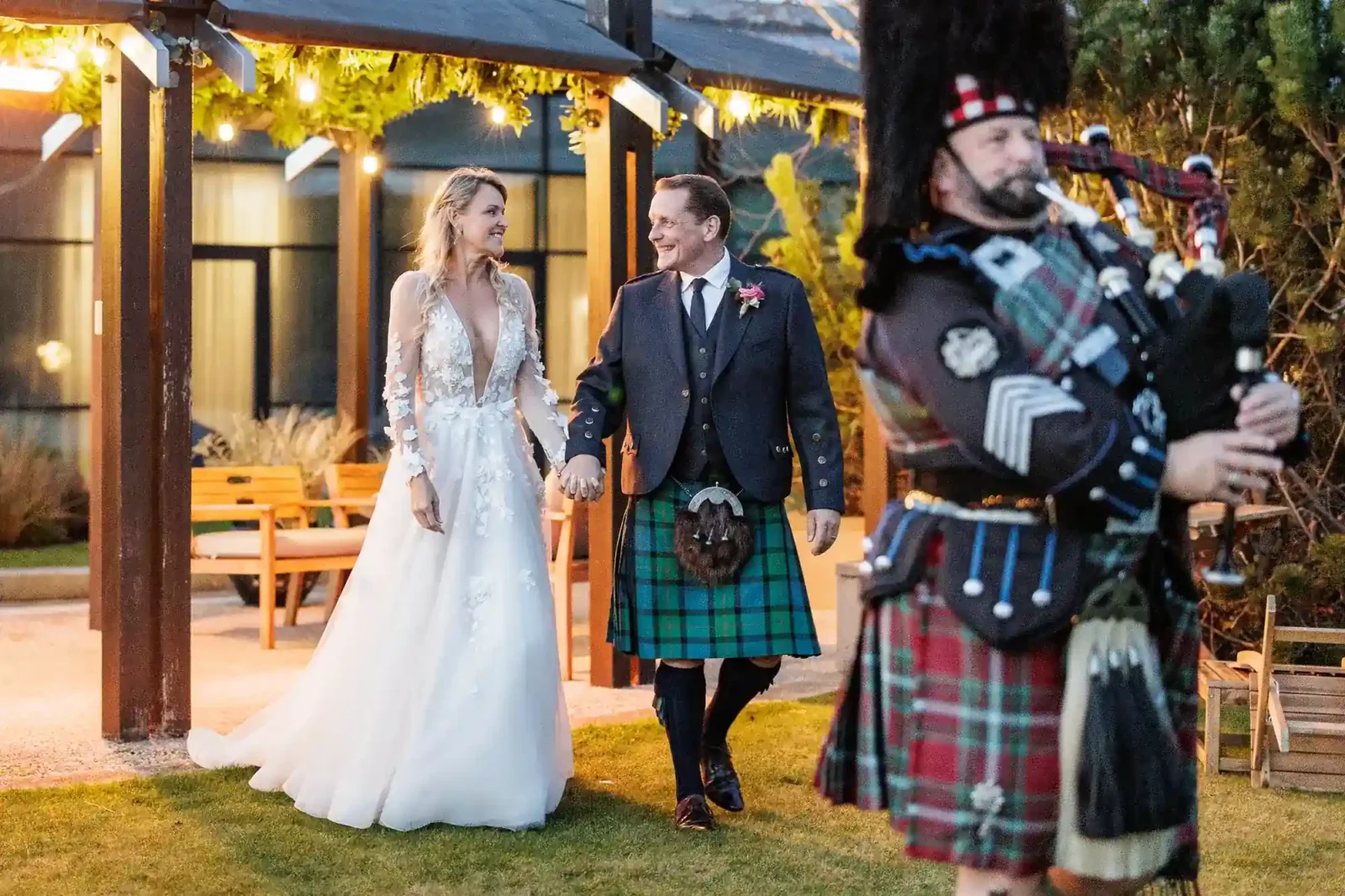 A bride in a white dress and a groom in a kilt hold hands, smiling at each other outdoors. Fairy lights hang above. A bagpiper in traditional Scottish attire plays in the foreground. Wooden benches and greenery are visible in the background of The Roof Garden at The Glasshouse Edinburgh.
