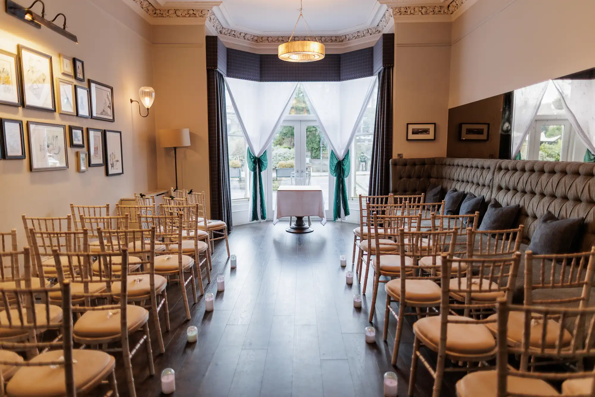 A wedding ceremony setup with rows of gold chairs facing a table in front of large bay windows with green draped curtains.