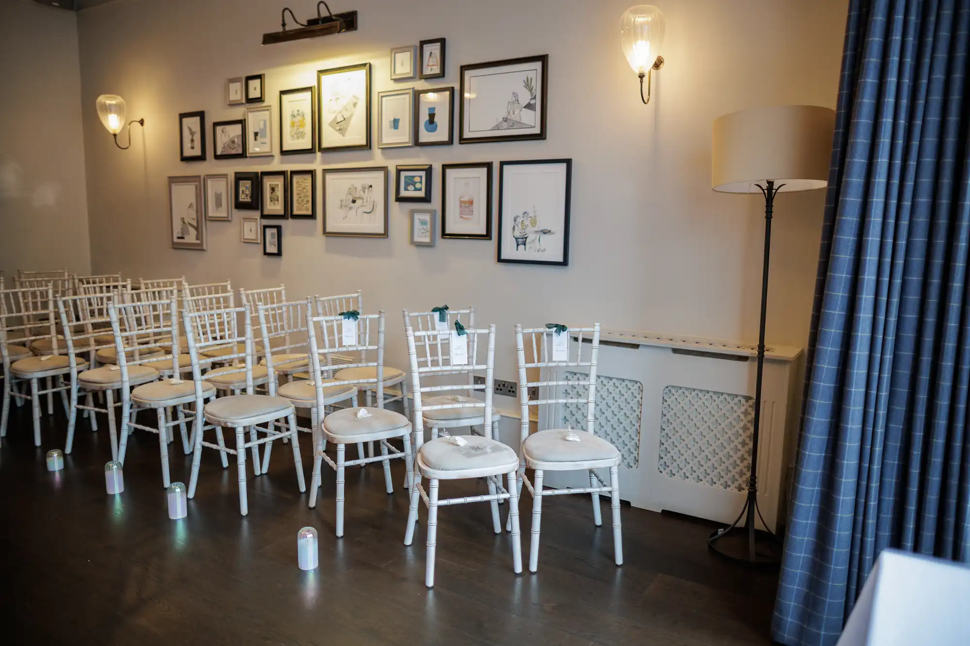 Rows of empty white chairs with light grey cushions are set up in an aisle, decorated with green ribbons. The wall behind them features framed artwork and a wall sconce.