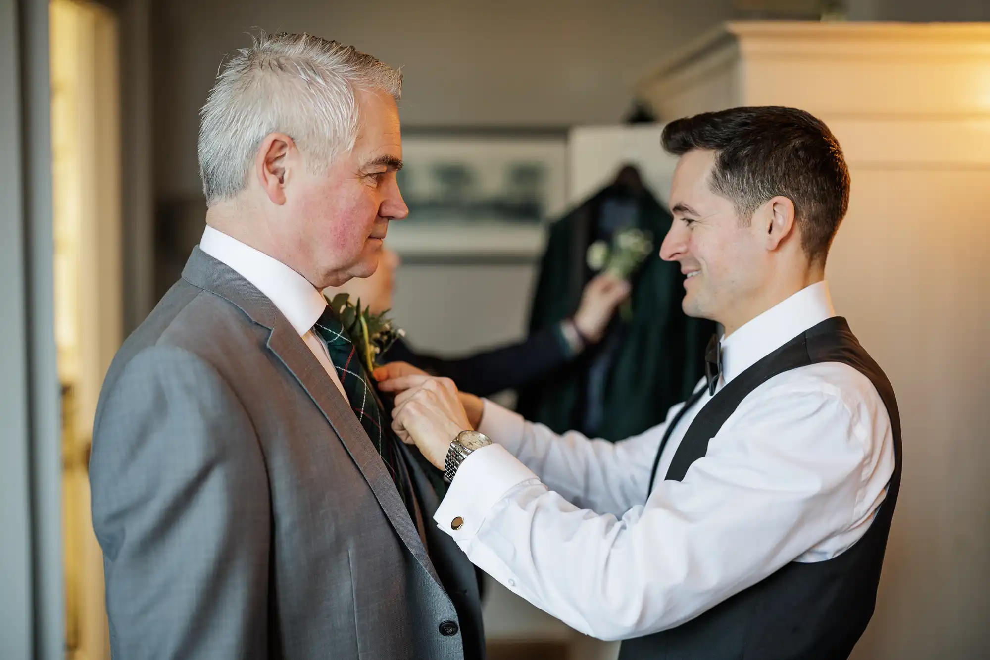 A groom in a white shirt, black waistcoat, and bow tie smiles as he pins a boutonniere onto the lapel of an older man's grey suit jacket. The older man has grey hair and is wearing a white shirt and green and red tartan tie.