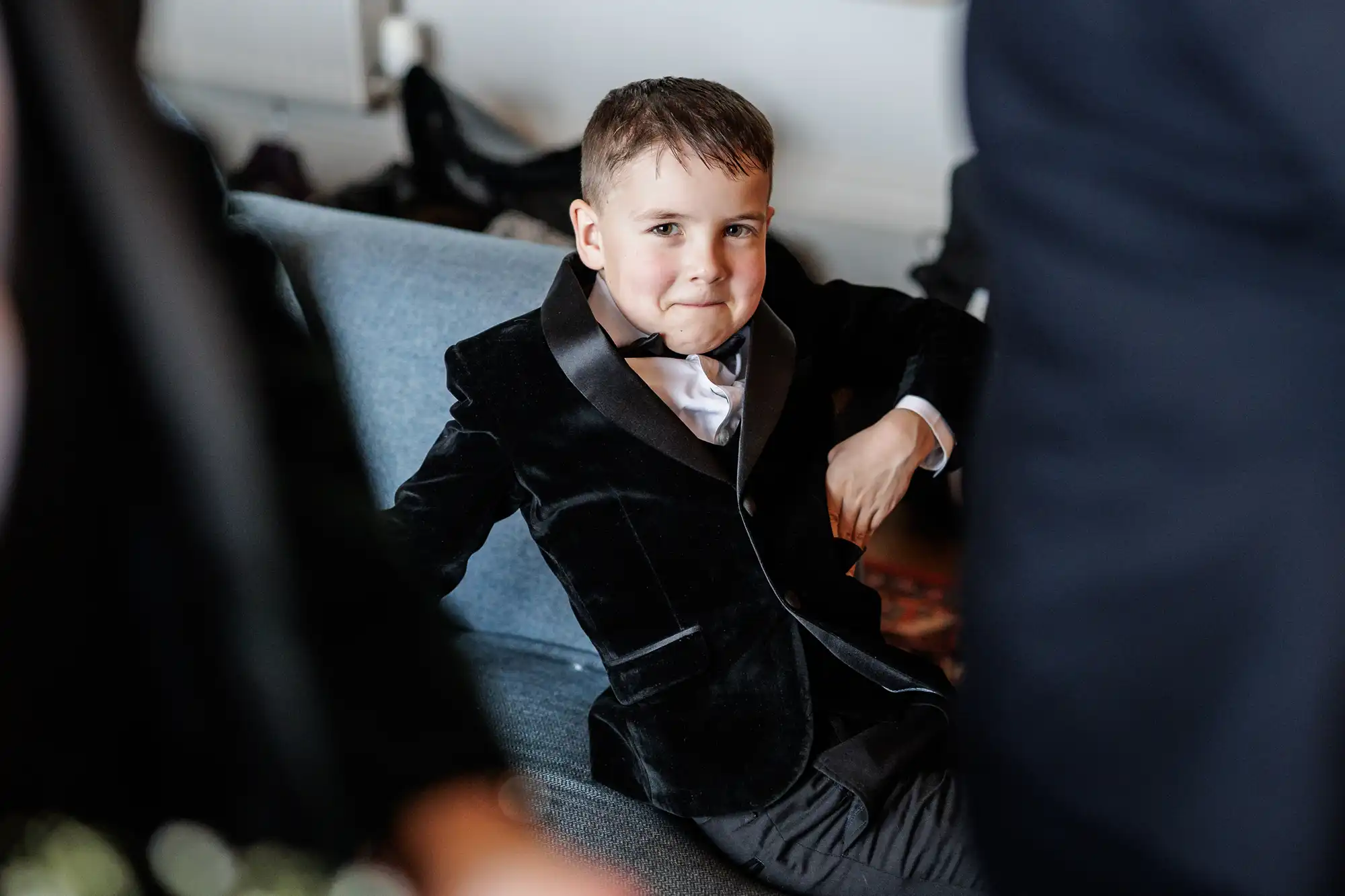 A young boy in a black velvet tuxedo with a bow tie sits on a sofa, looking at the camera.