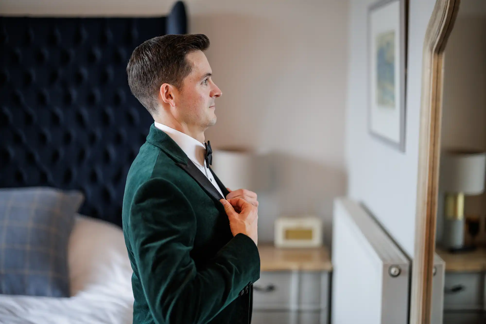 Man in a green velvet tuxedo adjusting lapel. He is looking into a mirror. Bedroom setting.