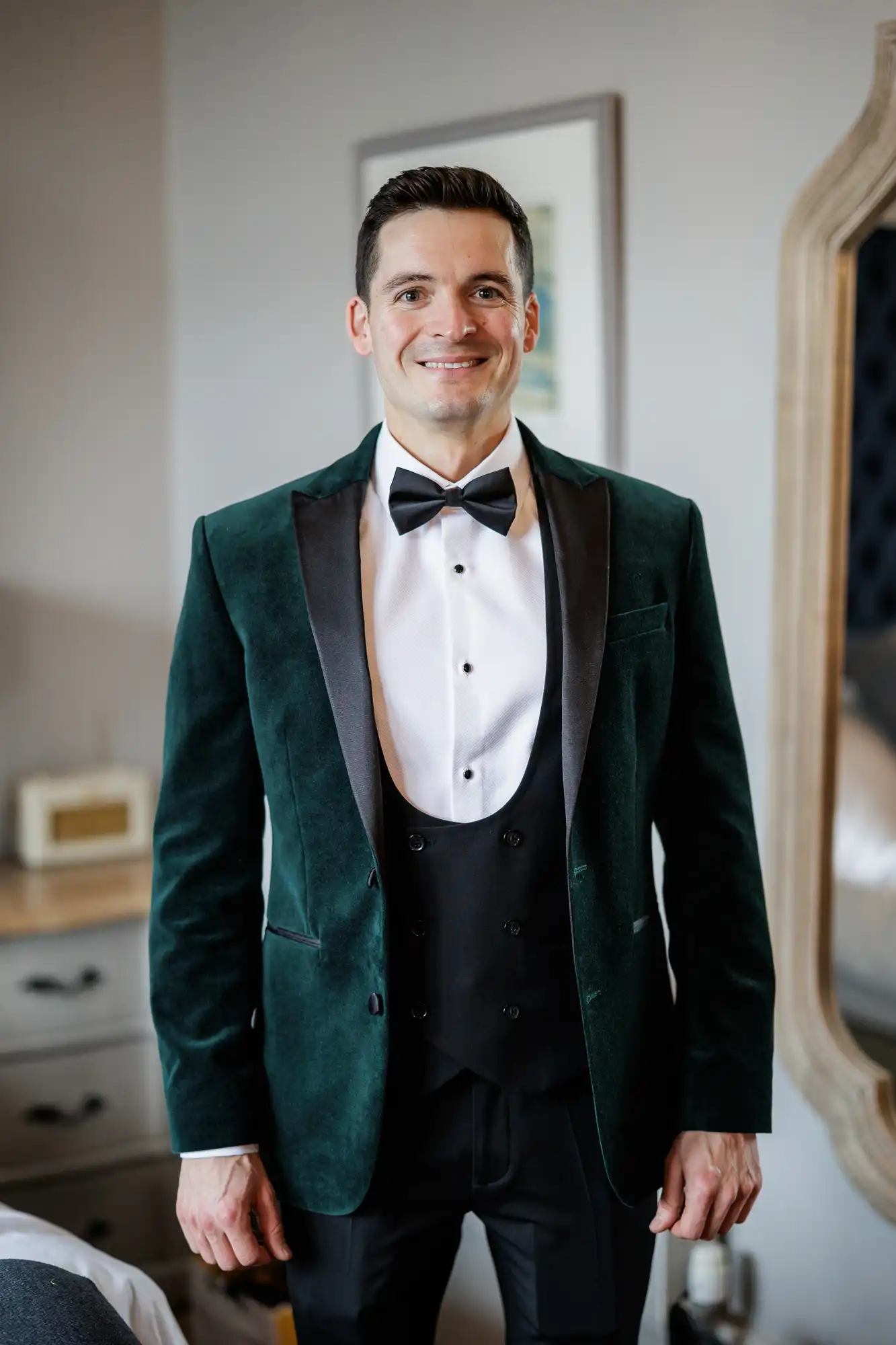 A man wears a dark green velvet tuxedo jacket with black lapels, a white formal shirt, black vest, and black bow tie. He smiles warmly.