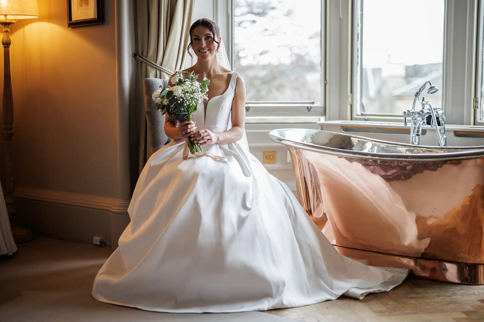 A smiling bride in a white gown sits holding a bouquet, next to a copper bathtub and a window.