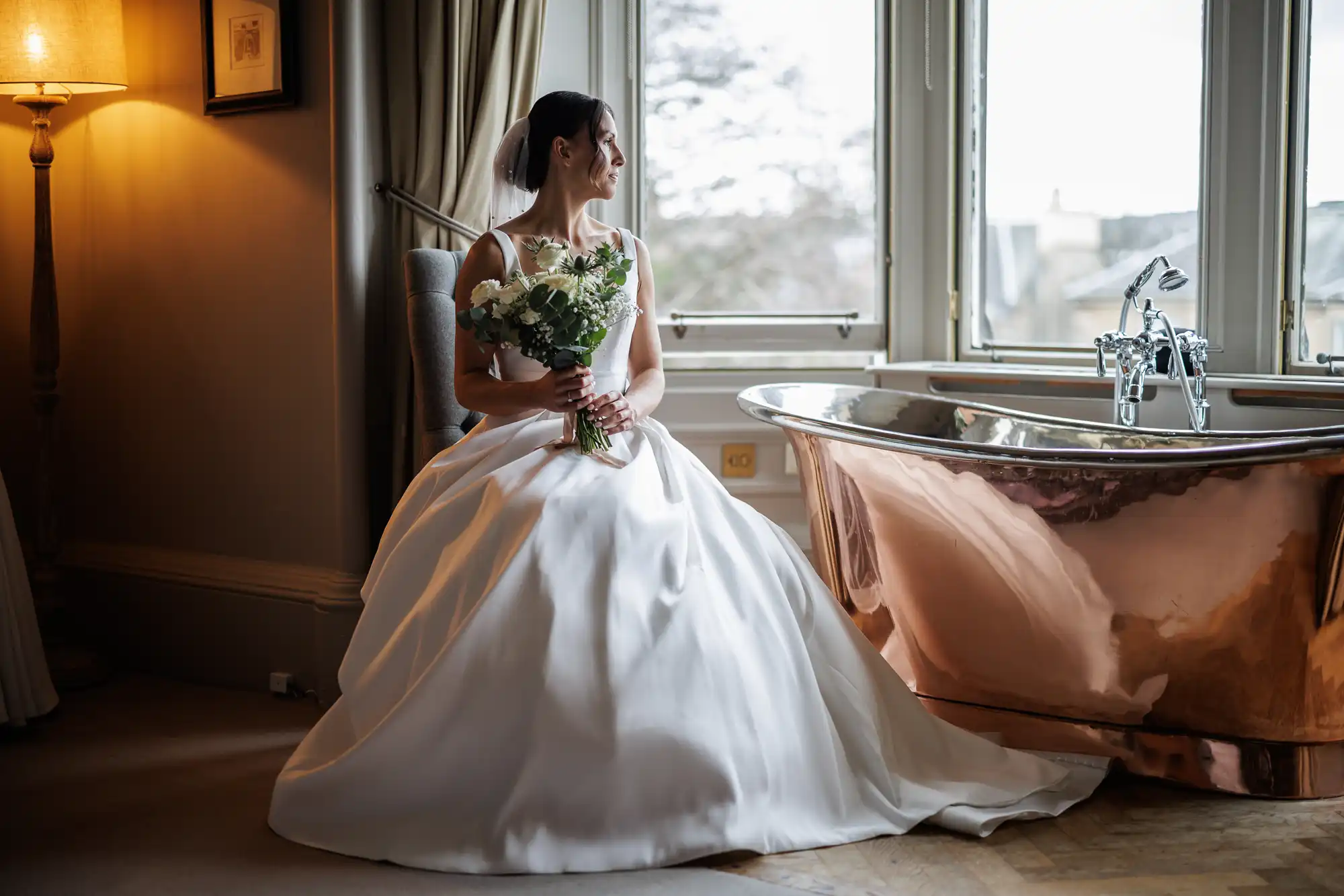 A bride in a white gown and veil sits holding a bouquet near a copper bathtub by a window.