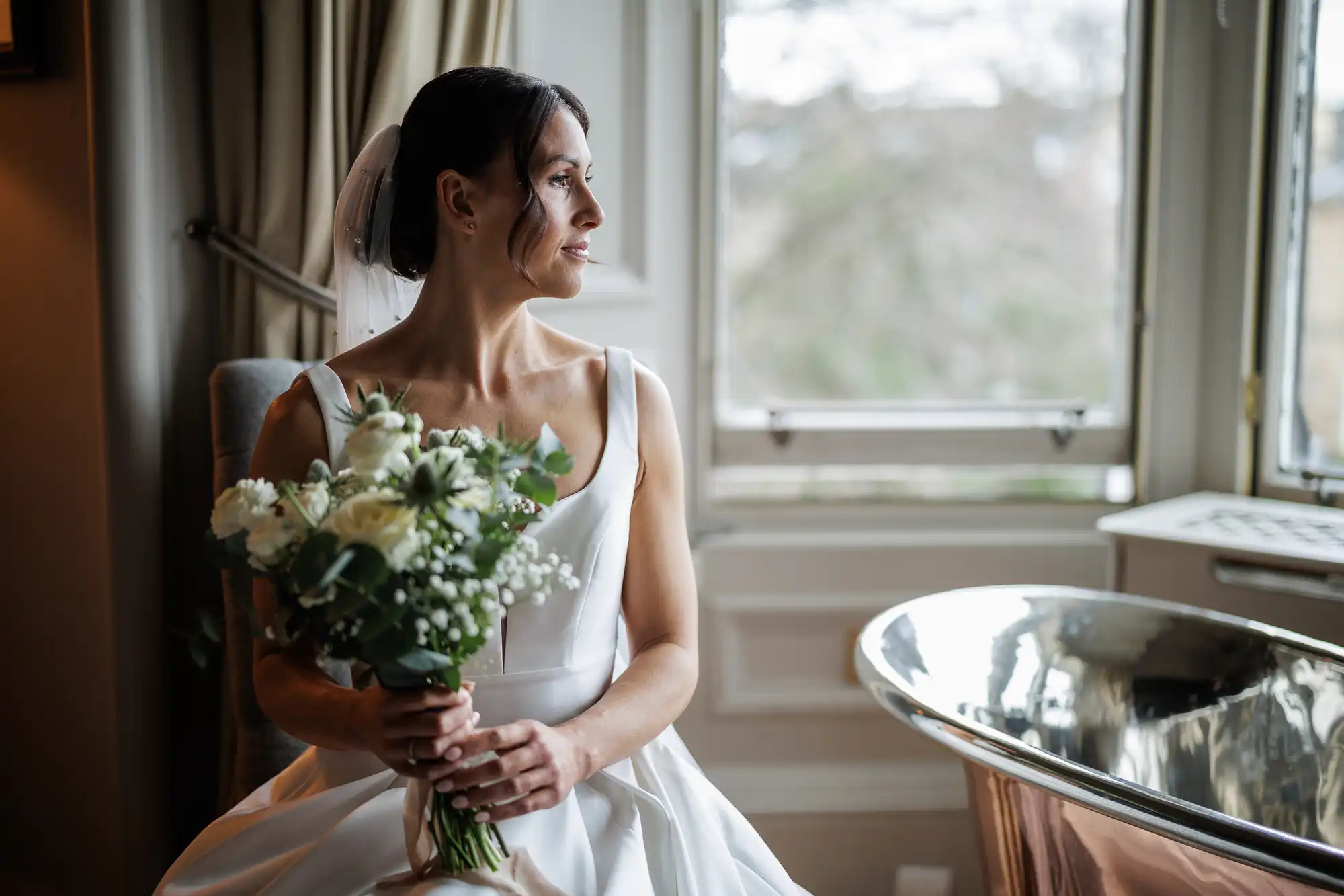 Bride in white dress and veil holds a bouquet of white roses and greenery, looking out a window. A copper bathtub is visible to the right.