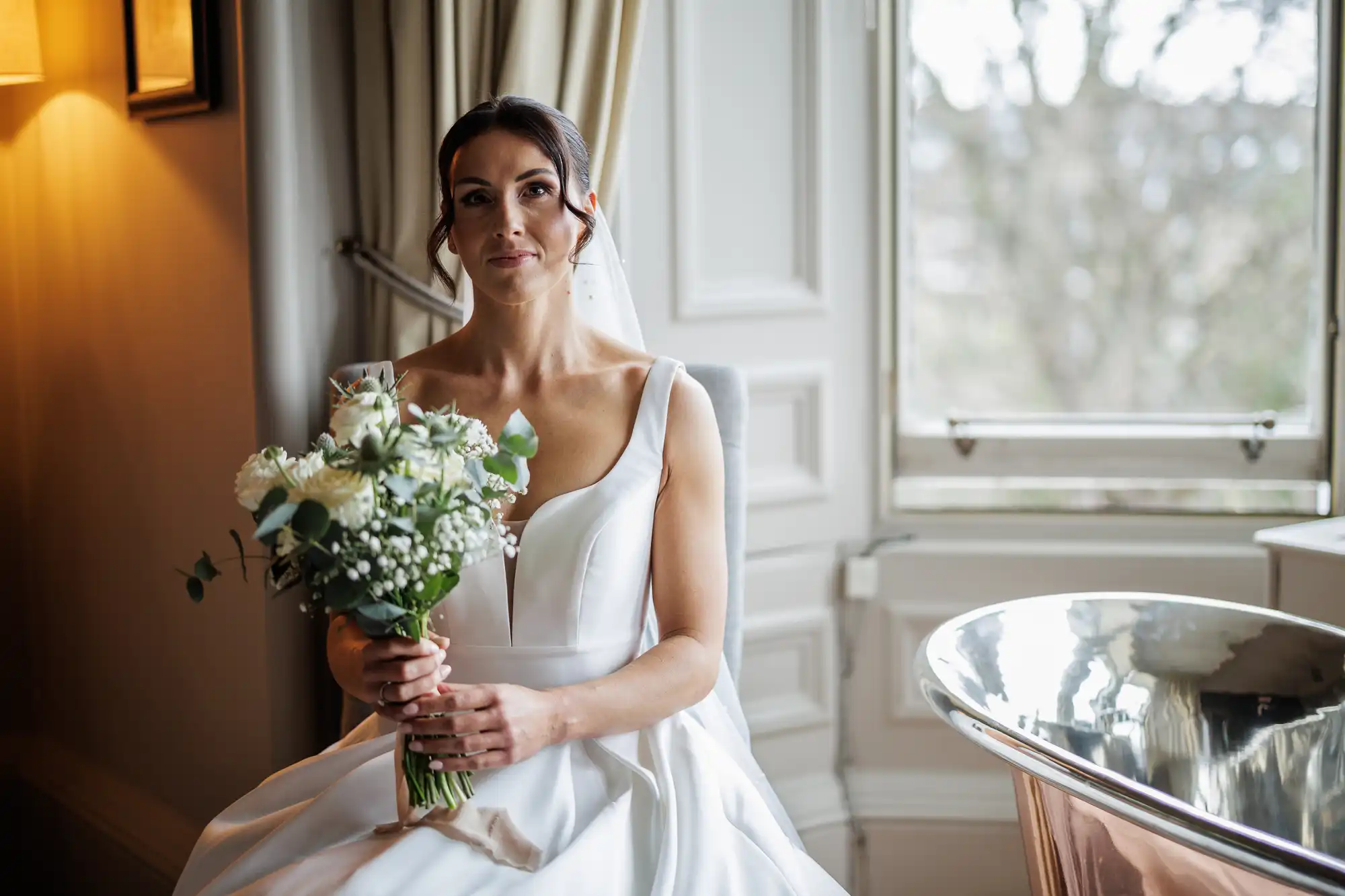A bride in a white satin gown holds a bouquet of white flowers and greenery, seated by a window with a copper bathtub in the foreground.