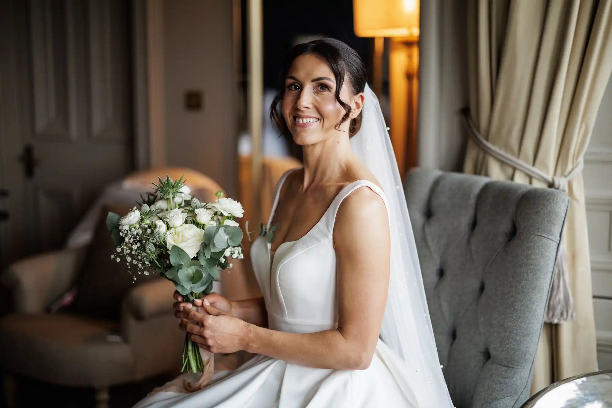 Bride in a white dress with veil, holding a white bouquet, sits in a chair, smiling.