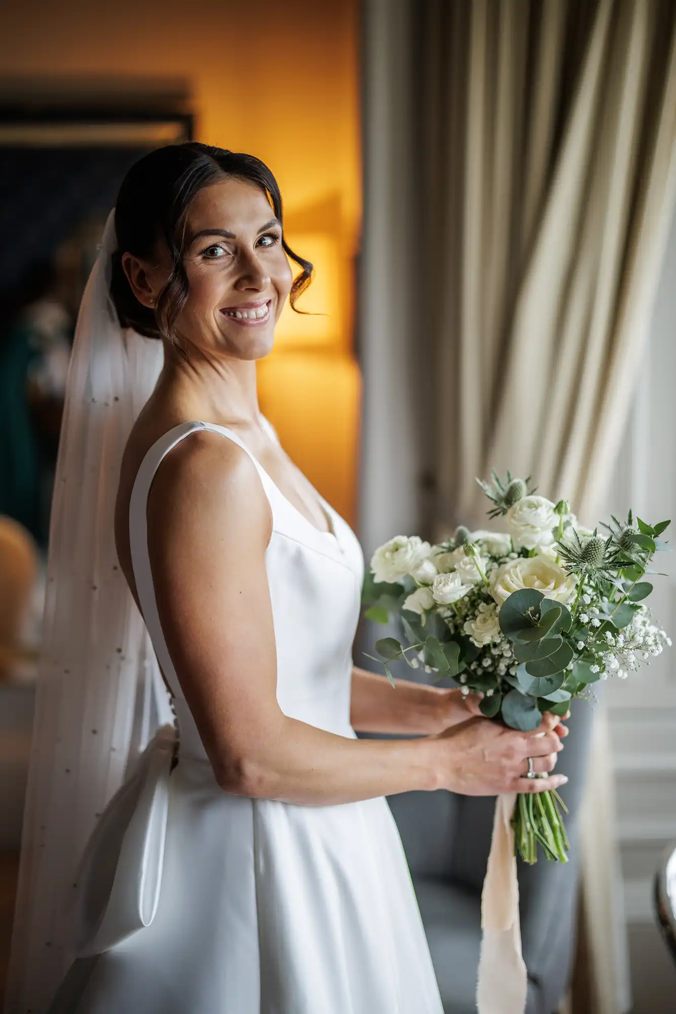 A smiling bride in a white dress and veil holds a bouquet of white roses, eucalyptus, and baby's breath.
