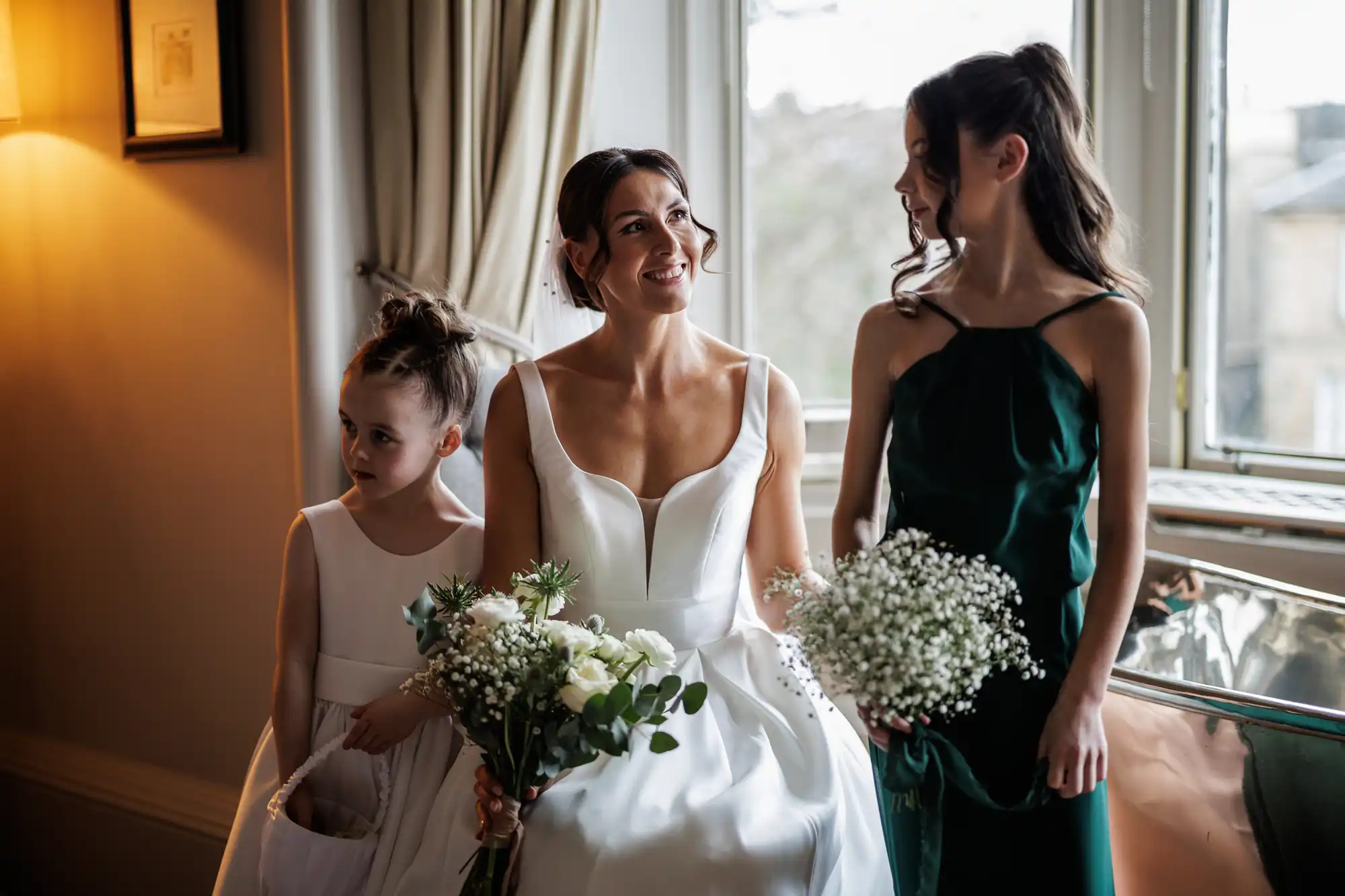 Bride in a white dress smiles at a younger girl in white holding a basket, and a bridesmaid in a green dress. They hold bouquets.
