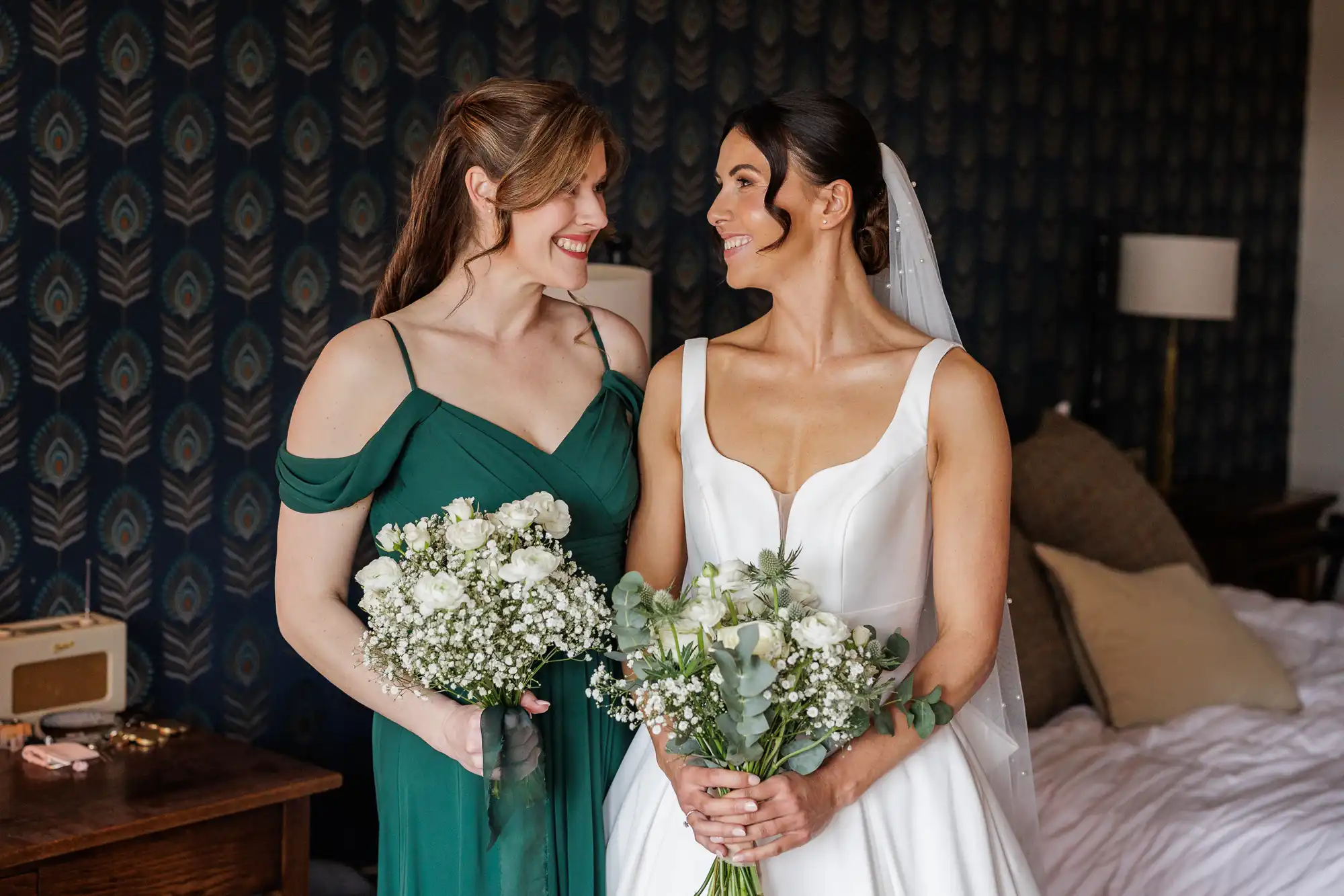 A bride in a white satin dress and her bridesmaid in an emerald green dress smile at each other, holding white bouquets.