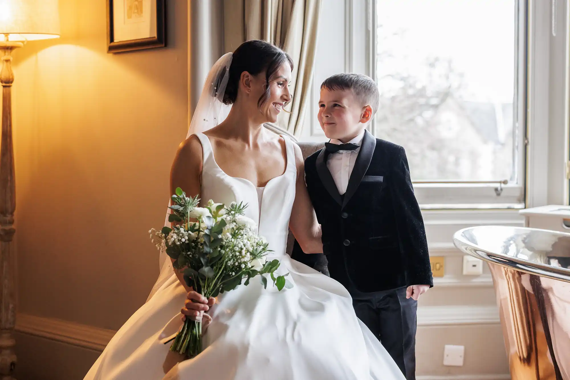 A bride in a white gown and veil smiles at a young boy in a tuxedo. They are holding hands and standing near a window.