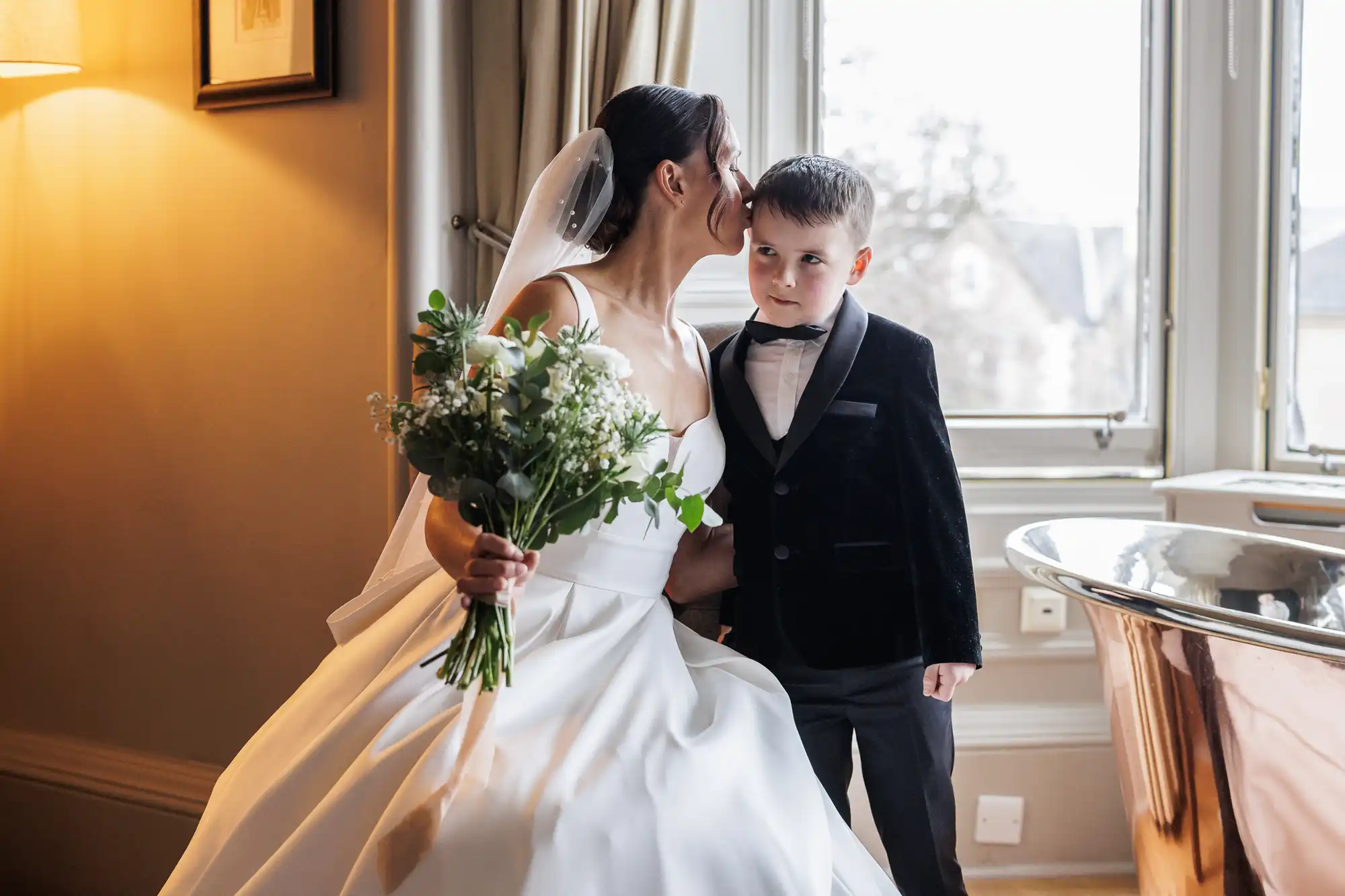 Bride in a white wedding dress and veil kisses a young boy in a black tuxedo on the forehead, holding a bouquet of white flowers.